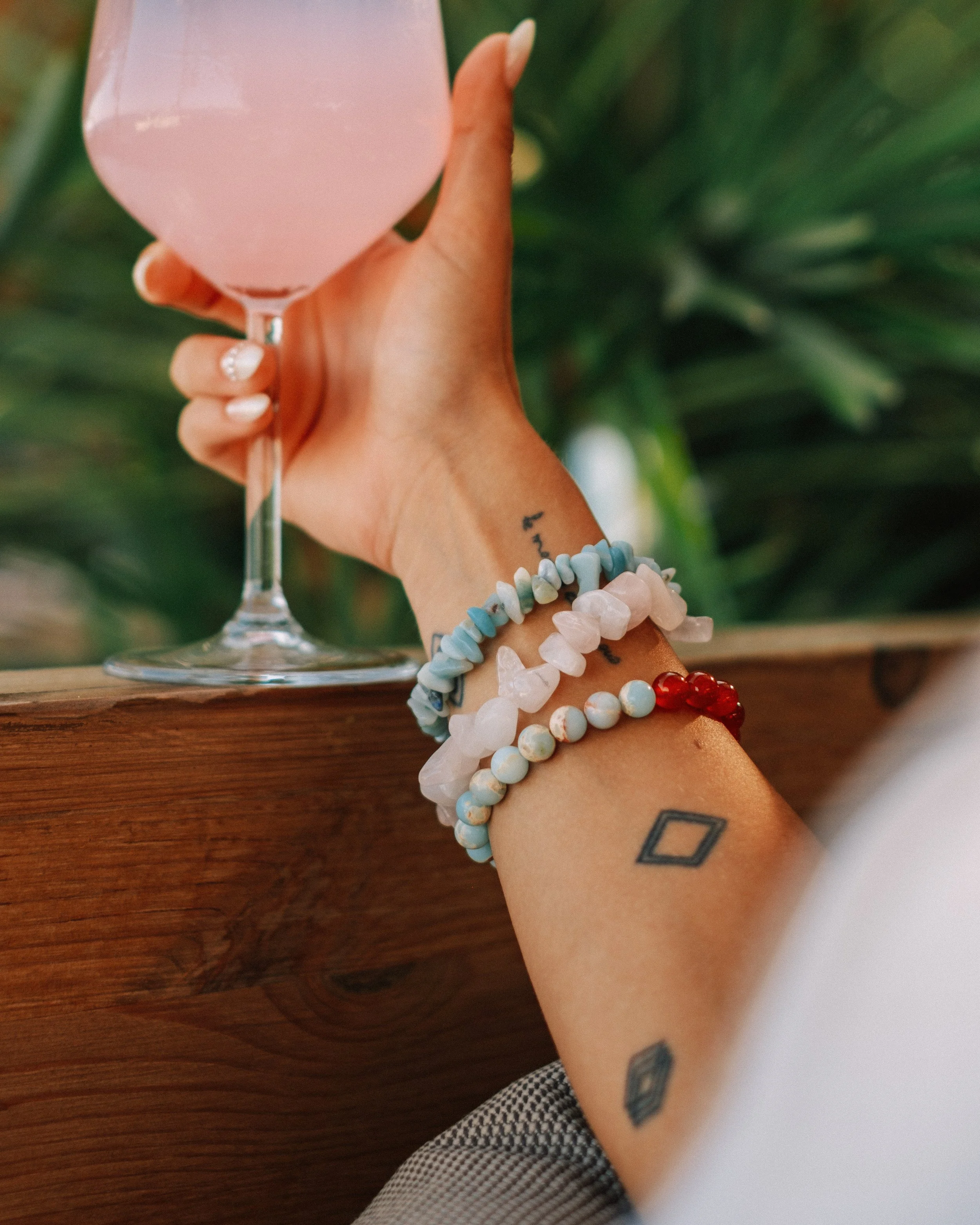 A person holding a pink drink in a wine glass, with their arm resting on a wooden surface, wearing multiple beaded bracelets and displaying tattoos on their arm. The background is green foliage.
