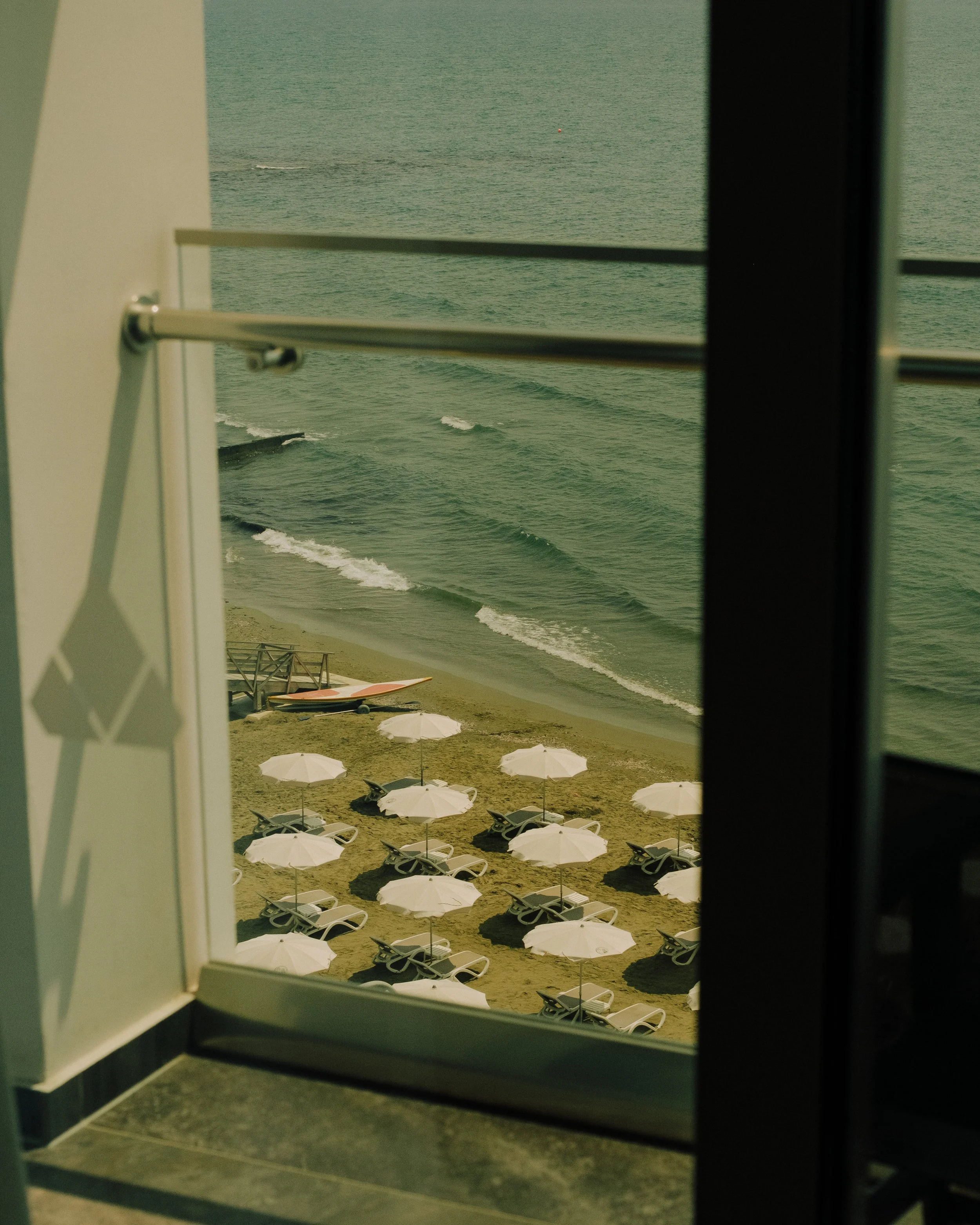 View of a beach with white umbrellas and lounge chairs, seen through a glass door with a railing outside.