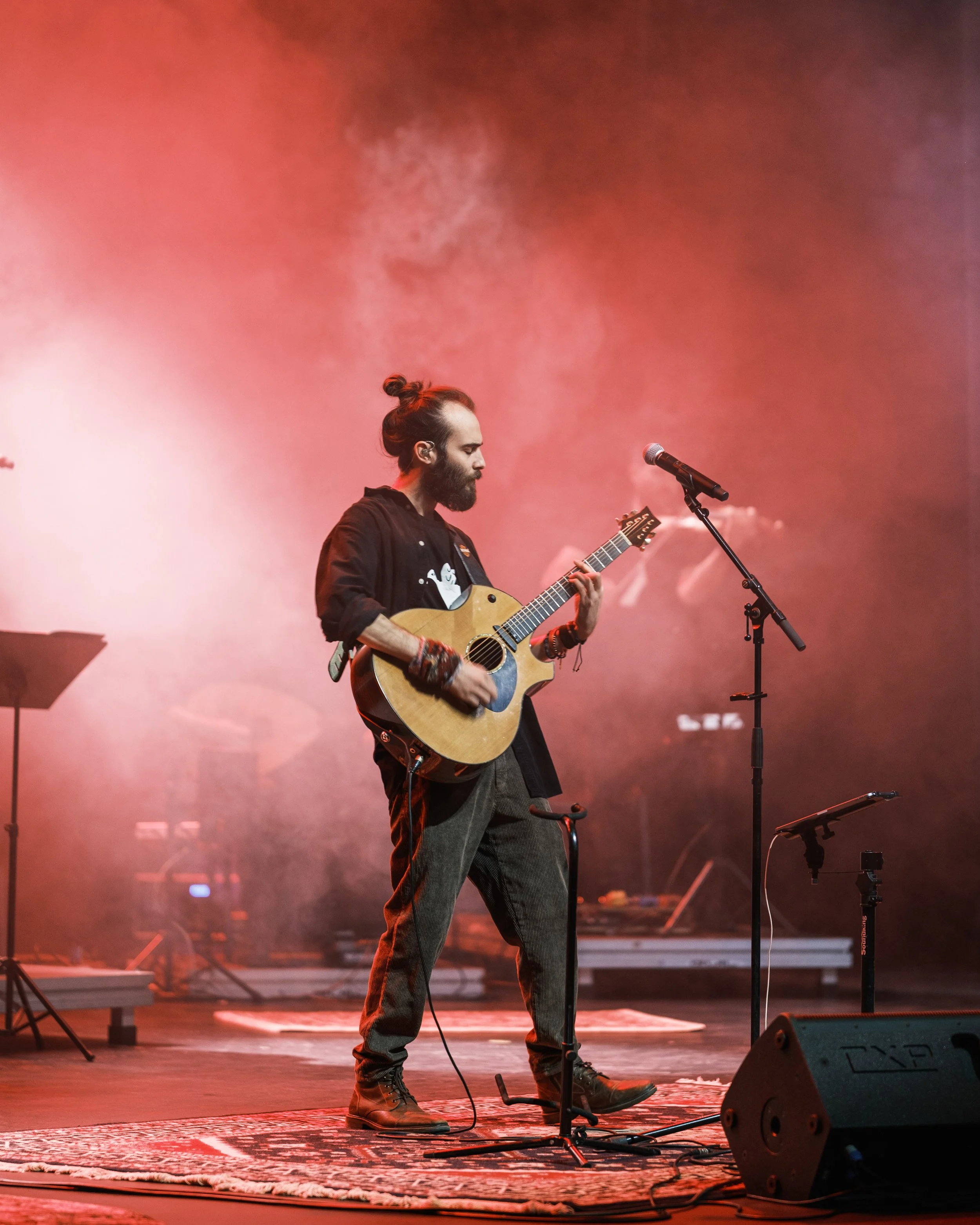 A man with a beard and bun hairstyle playing an acoustic guitar on stage with pink and orange lighting, standing on a rug, with microphone and stage equipment around.
