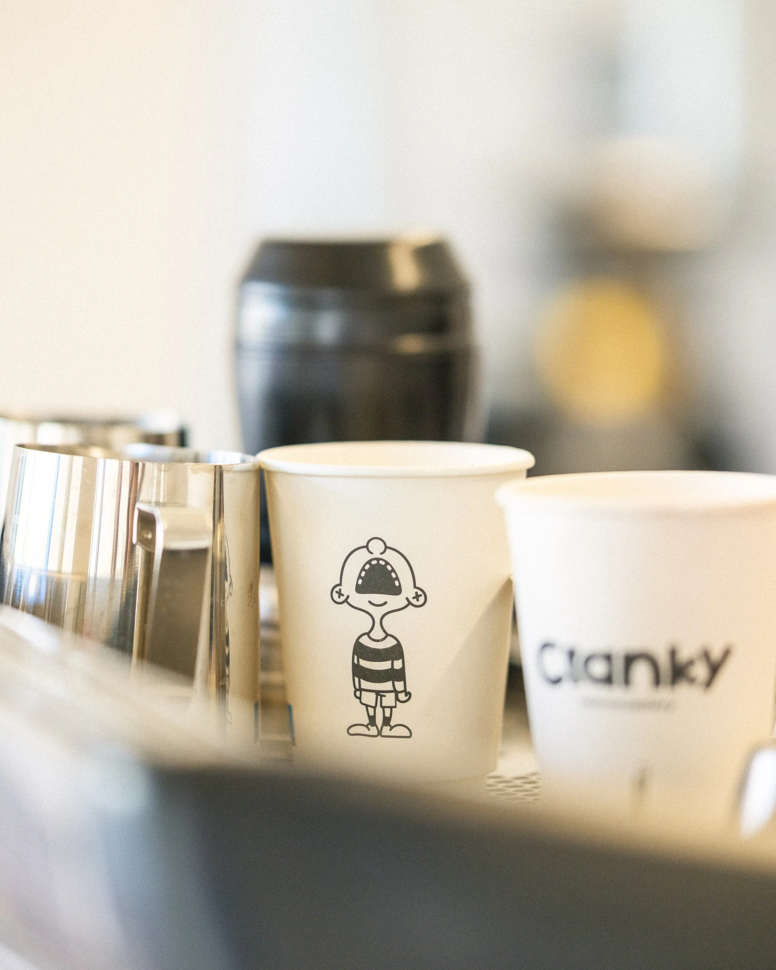 Close-up of two white paper cups with cartoon characters and the word 'Cranky' on one cup, with blurred coffee machine in the background.
