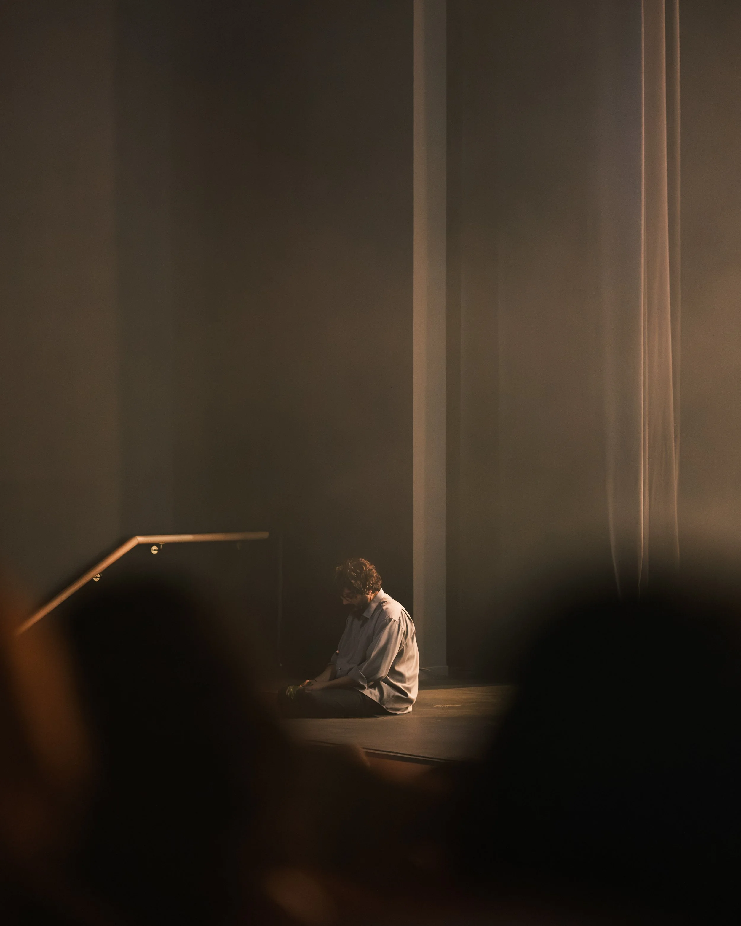Person sitting alone on stage, head bowed, with dark background and focused lighting.