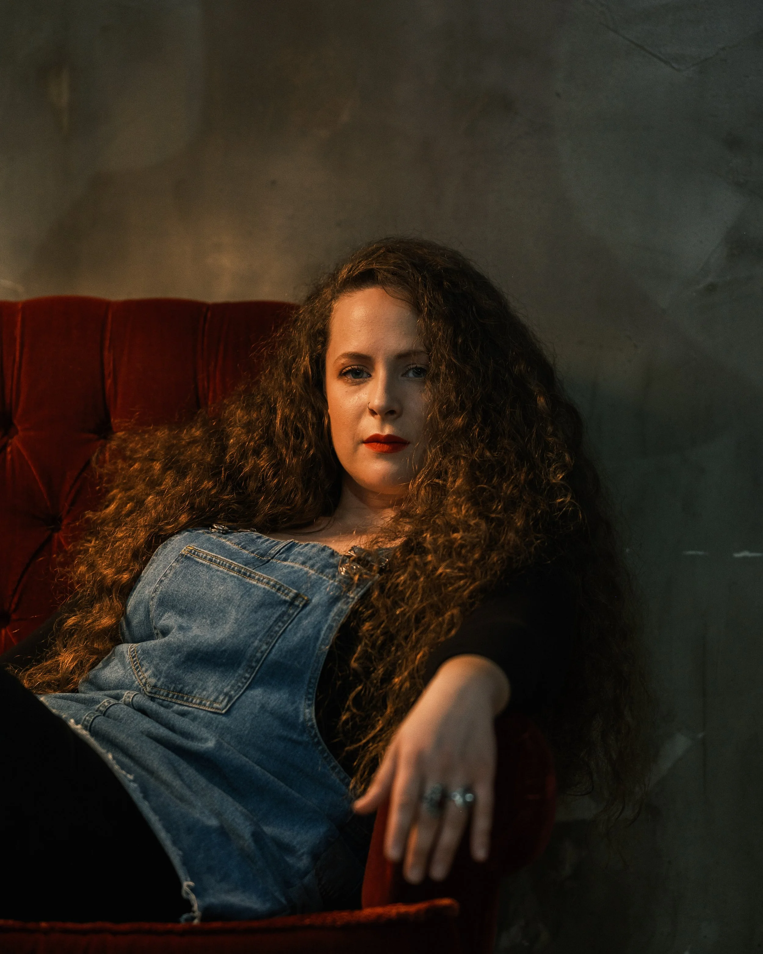 A woman with long curly hair lounging on a red velvet sofa, wearing a black top and denim overalls, looking at the camera with a relaxed expression.