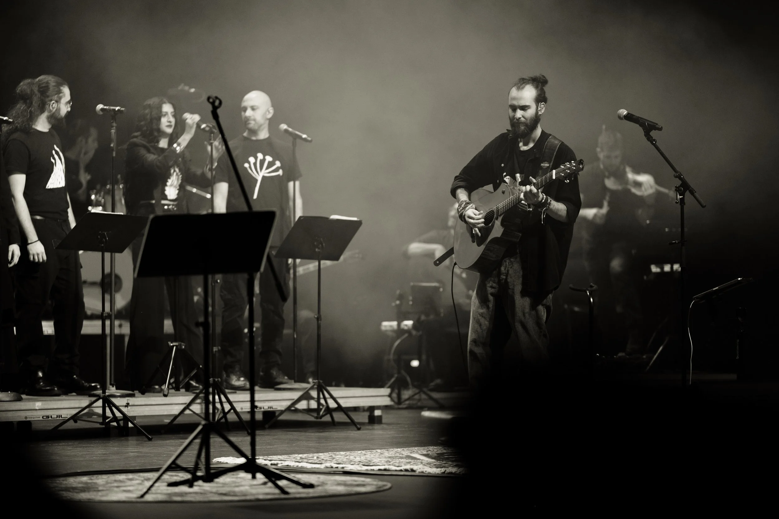 A black and white photo of a music performance on stage. On the right, a man with a beard and bun is playing an acoustic guitar. On the left, five women are singing into microphones, with music stands in front of them. The stage is dimly lit with vis