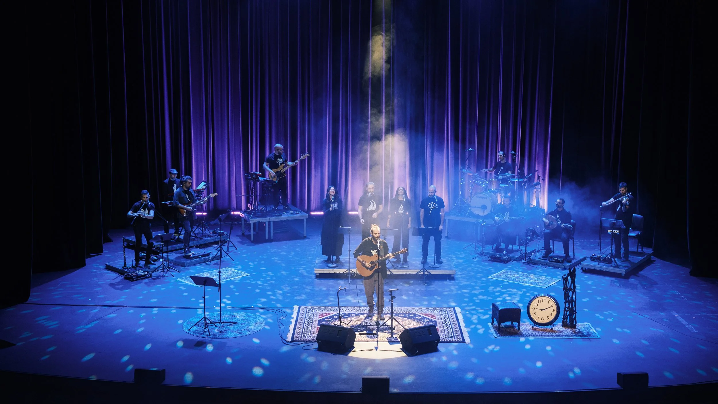 A live performance on a stage with multiple musicians, including a person playing guitar and singing in the center, with others playing keyboards, drums, bass guitar, and wind instruments. The stage is decorated with rugs, a clock, and various musica