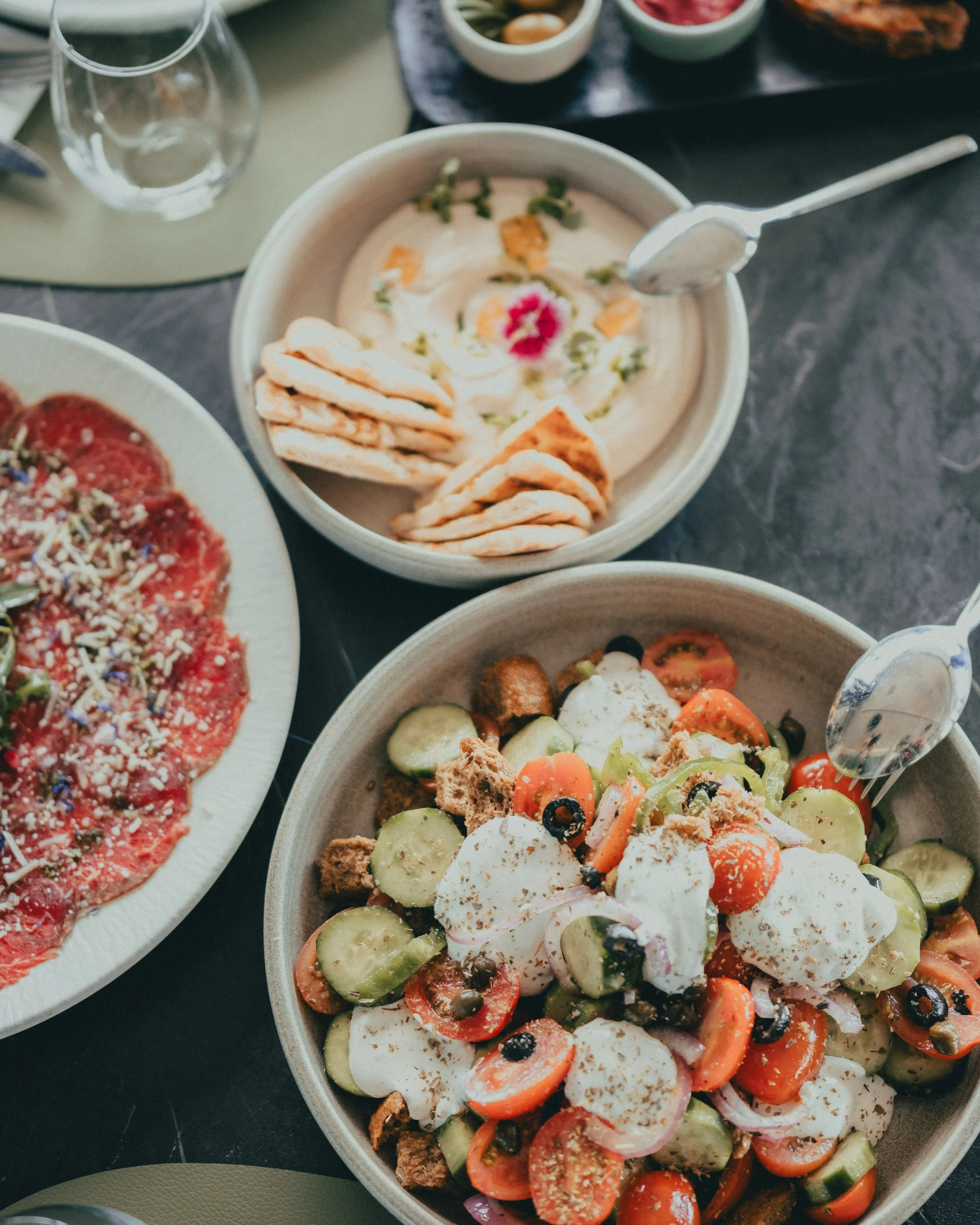 A Mediterranean meal featuring a fresh vegetable salad with tomatoes, cucumbers, olives, mozzarella, and croutons in a bowl; a plate with thinly sliced carpaccio topped with herbs and Parmesan cheese; and a bowl of creamy hummus garnished with olive 