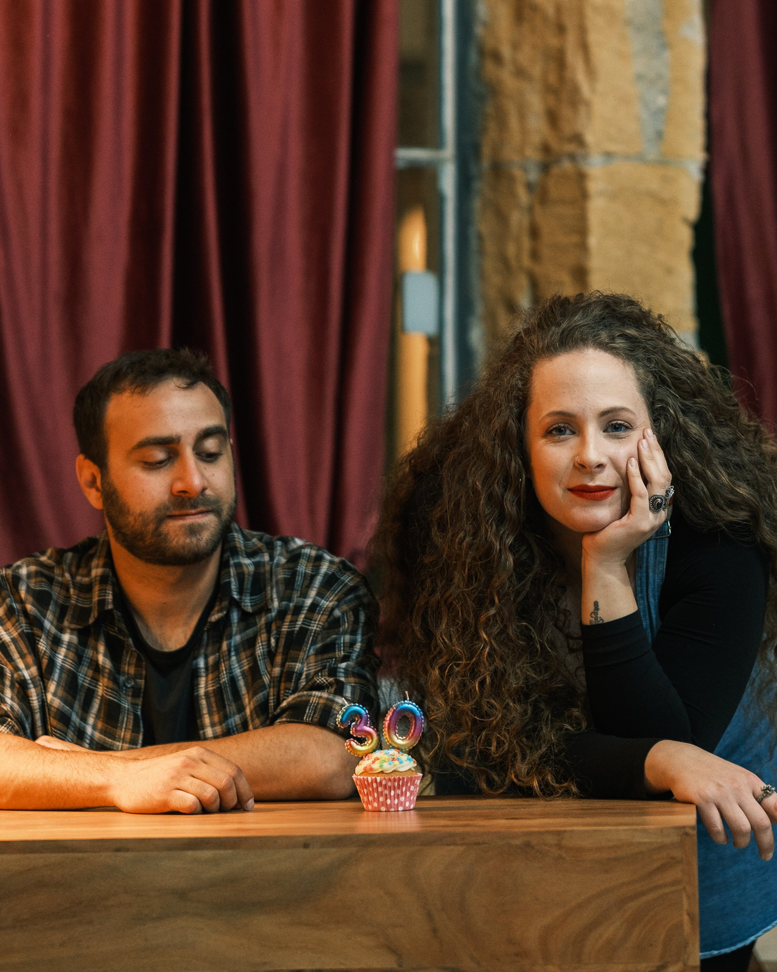 Two people sitting at a table with a cupcake that has a candle with the number 30, in a room with red curtains and brick wall.