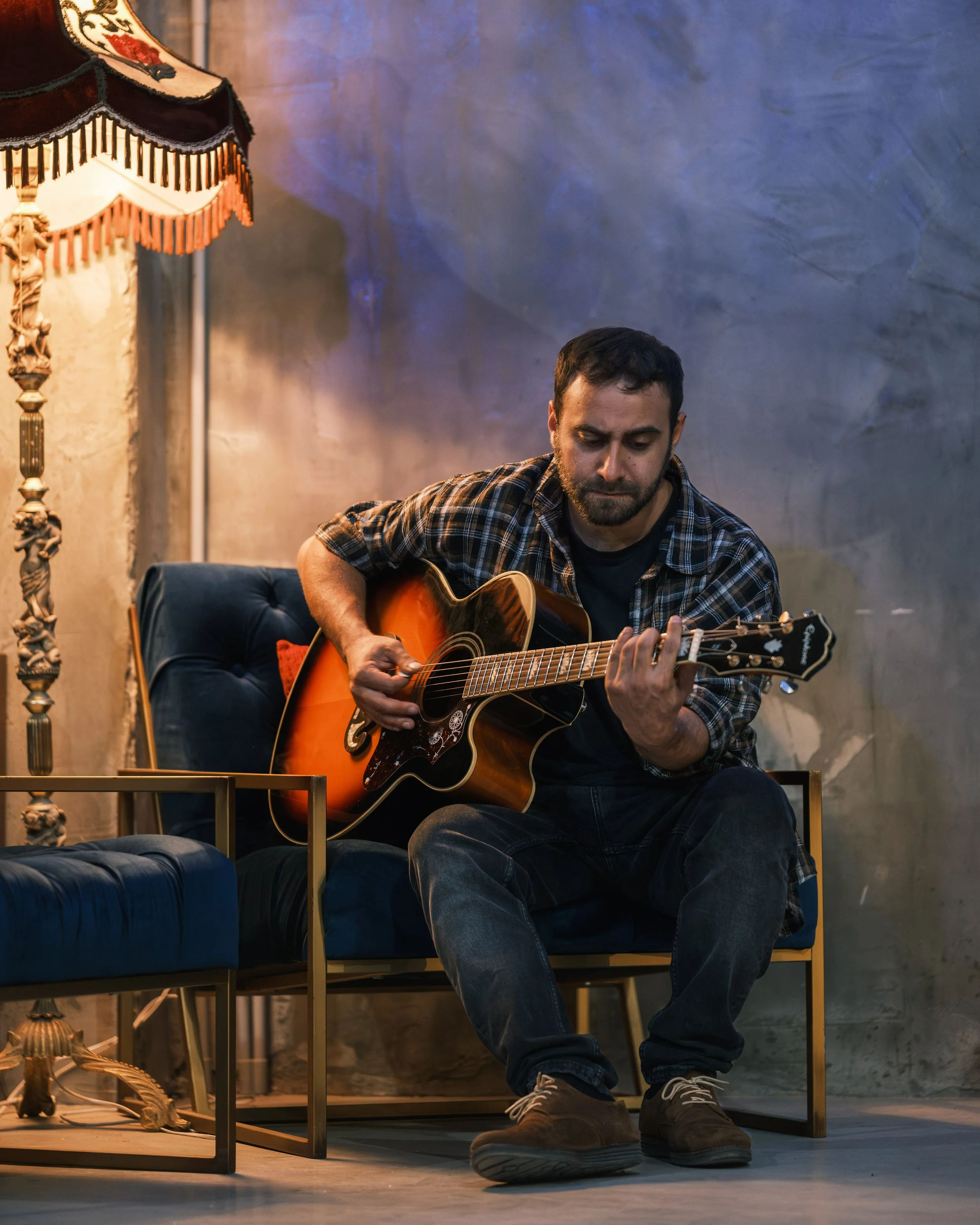 A man sitting on a dark blue sofa playing an acoustic guitar in a cozy room with a vintage lamp and textured wall.