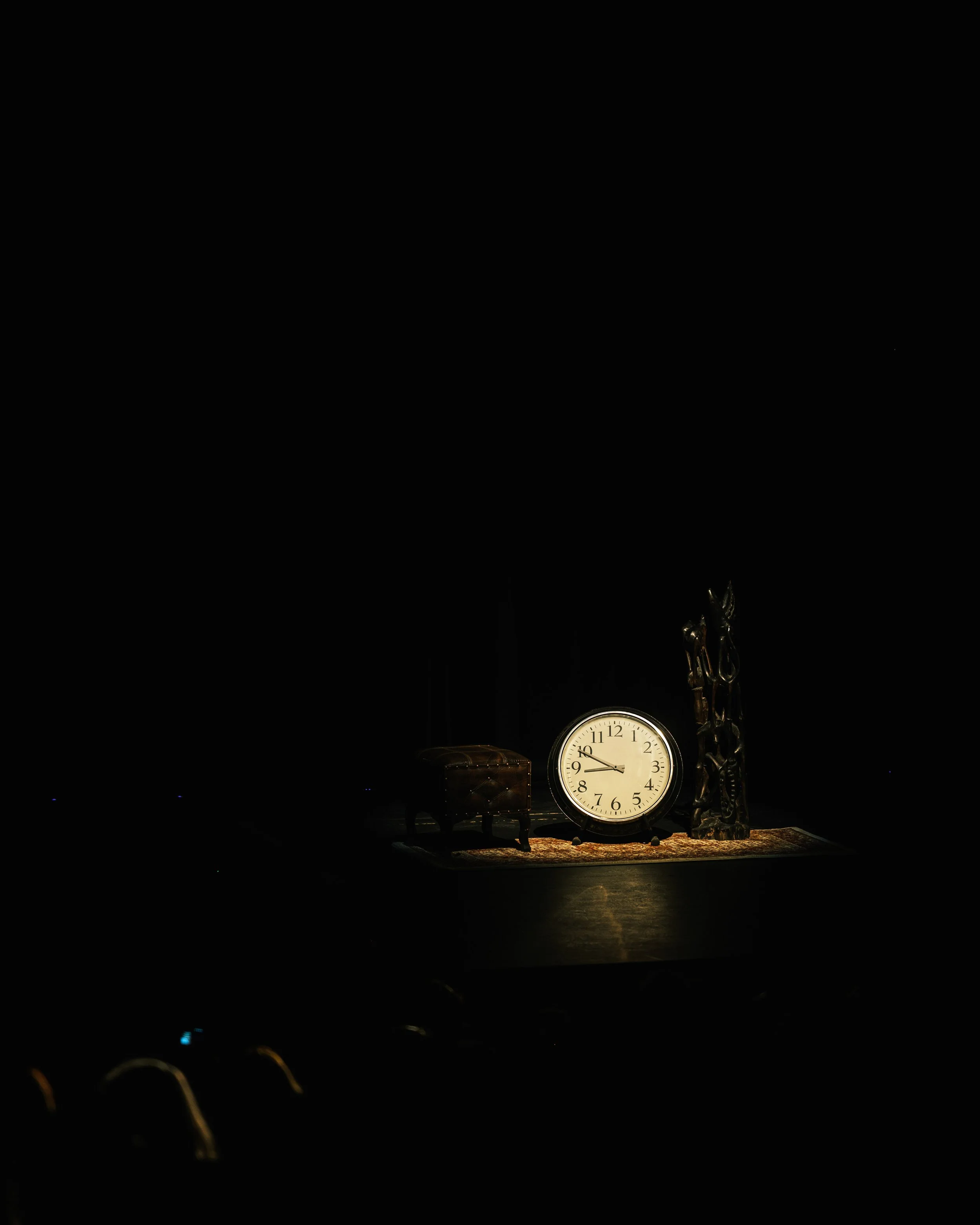 A stage with a standing lamp, a small stool, and a wall sculpture, illuminated by a single spotlight in a dark theater or auditorium.