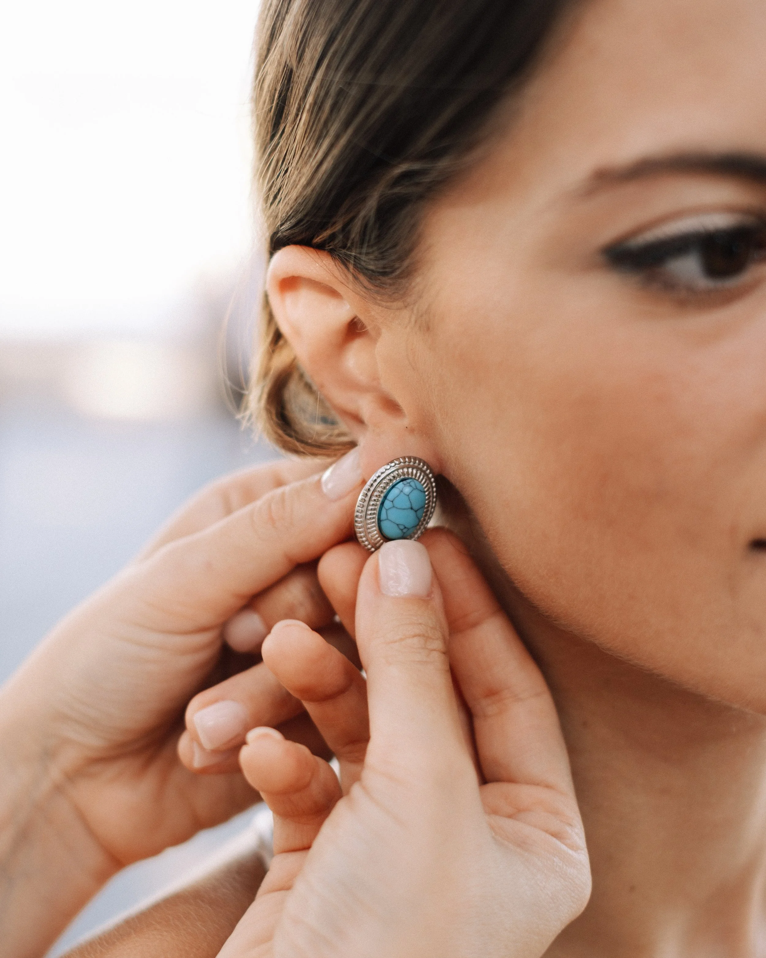 A woman is wearing a turquoise gemstone earring, which is being adjusted by another person's hand.