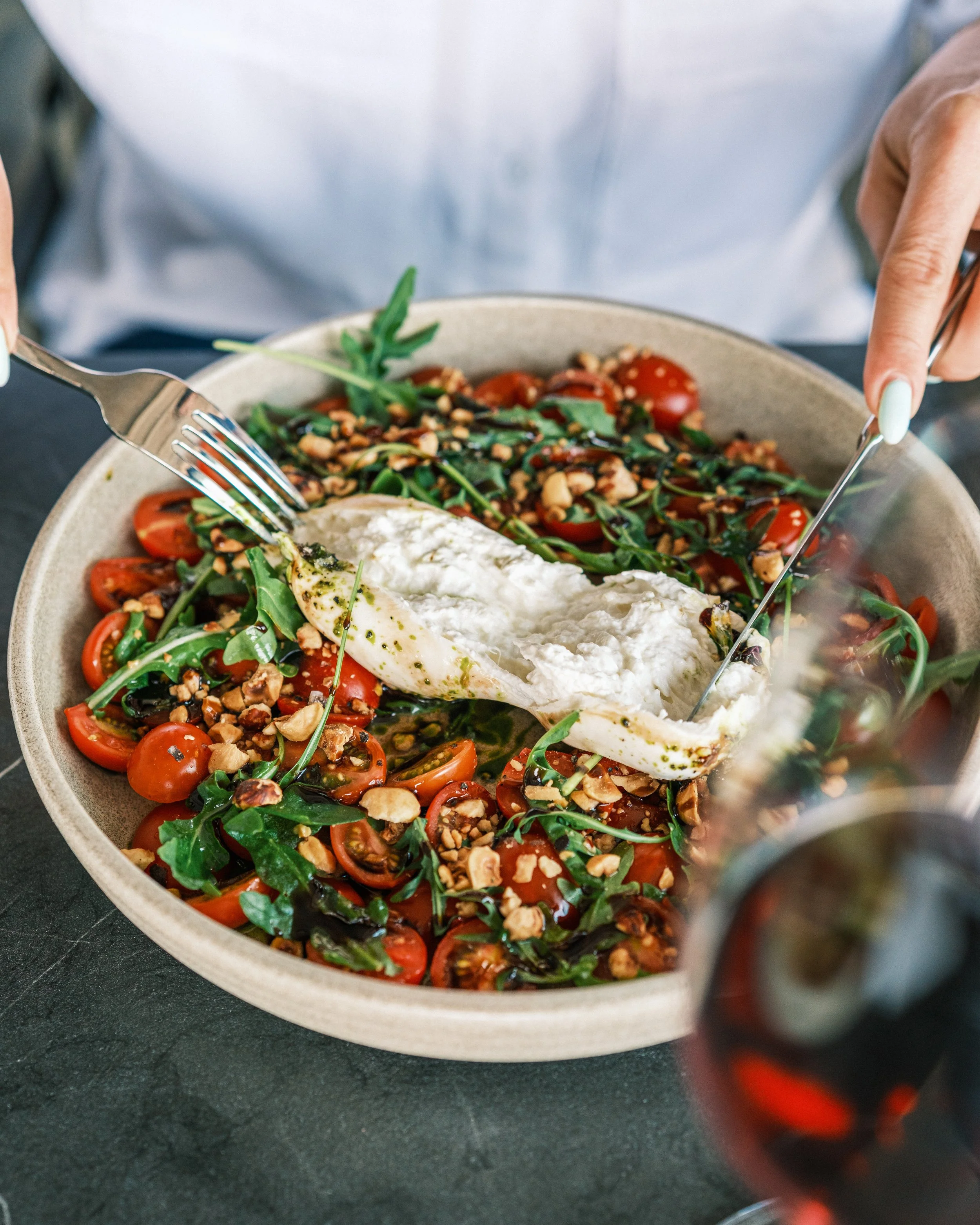 Person serving a fresh tomato and arugula salad with chopped nuts and a dollop of burrata cheese, accompanied by a glass of red wine.