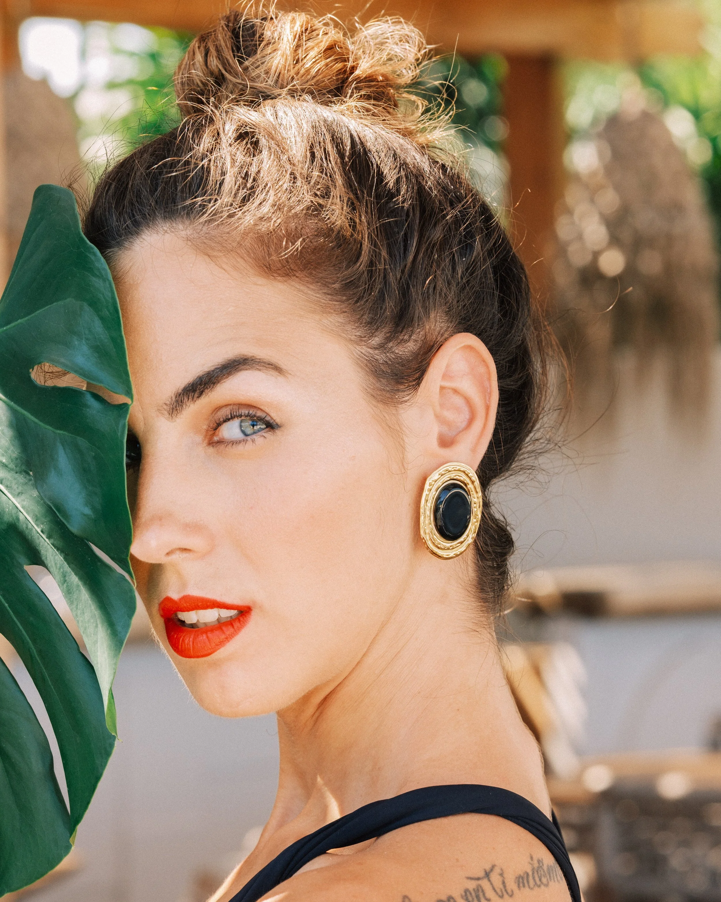 Close-up of a woman with styled brown hair, blue eyes, wearing red lipstick, gold and black earrings, and a black top, partially obscured by a large green leaf, outdoors with blurred background.