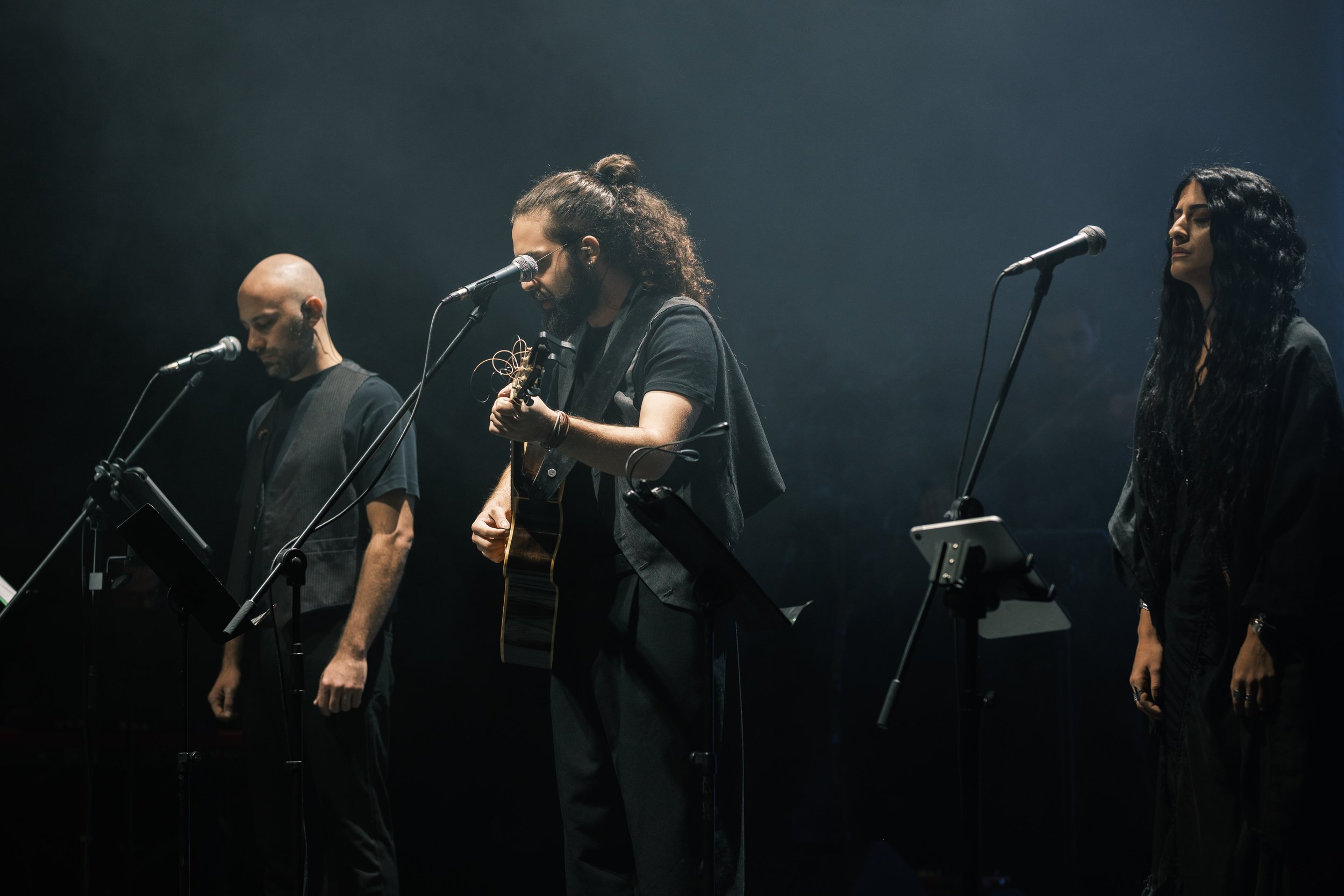 Three musicians standing on stage with microphones, one playing guitar, in a dimly lit performance setting.