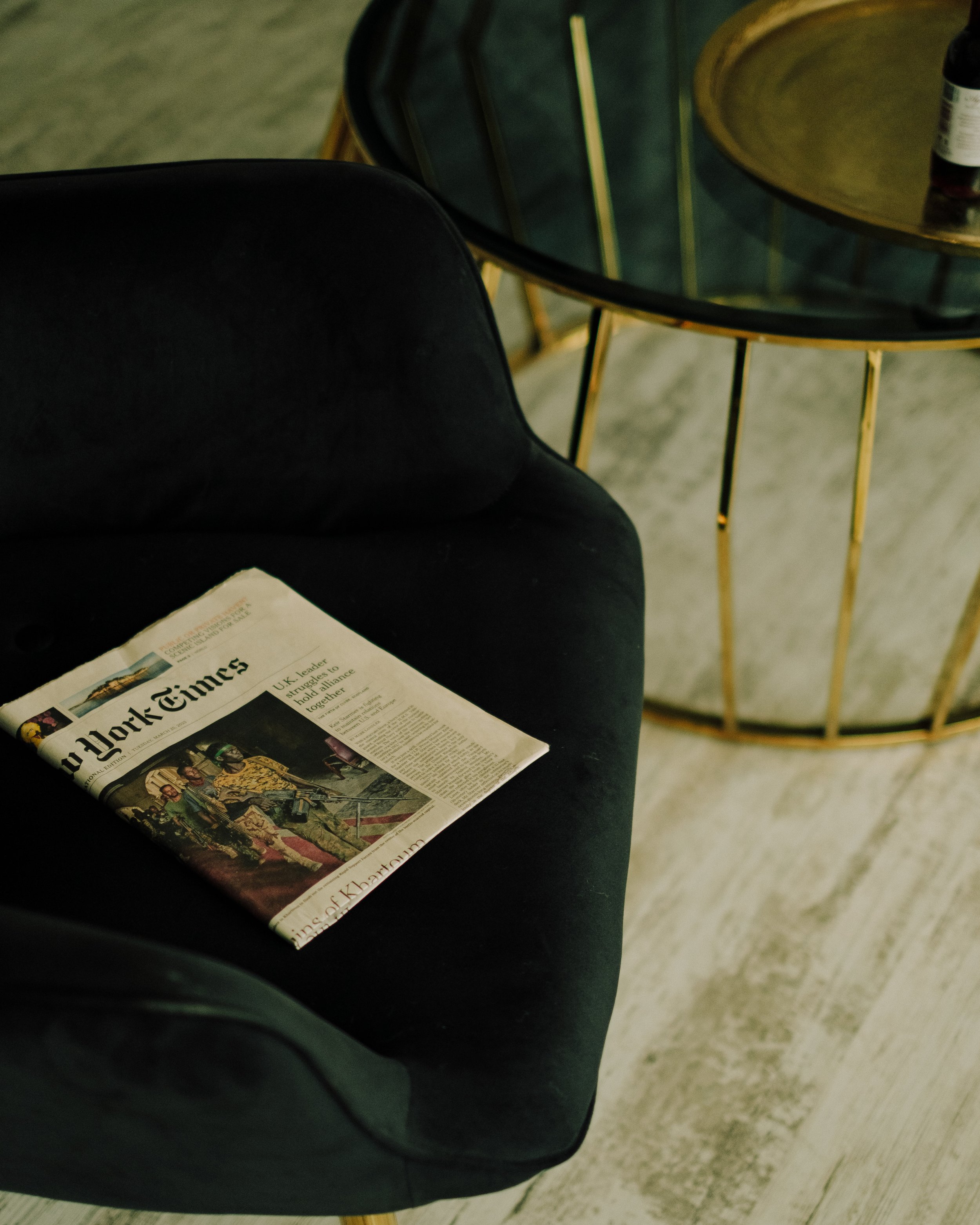 A black chair with a United States newspaper resting on it, and a round side table with a glass top and metal frame in a room with light-colored wood flooring.