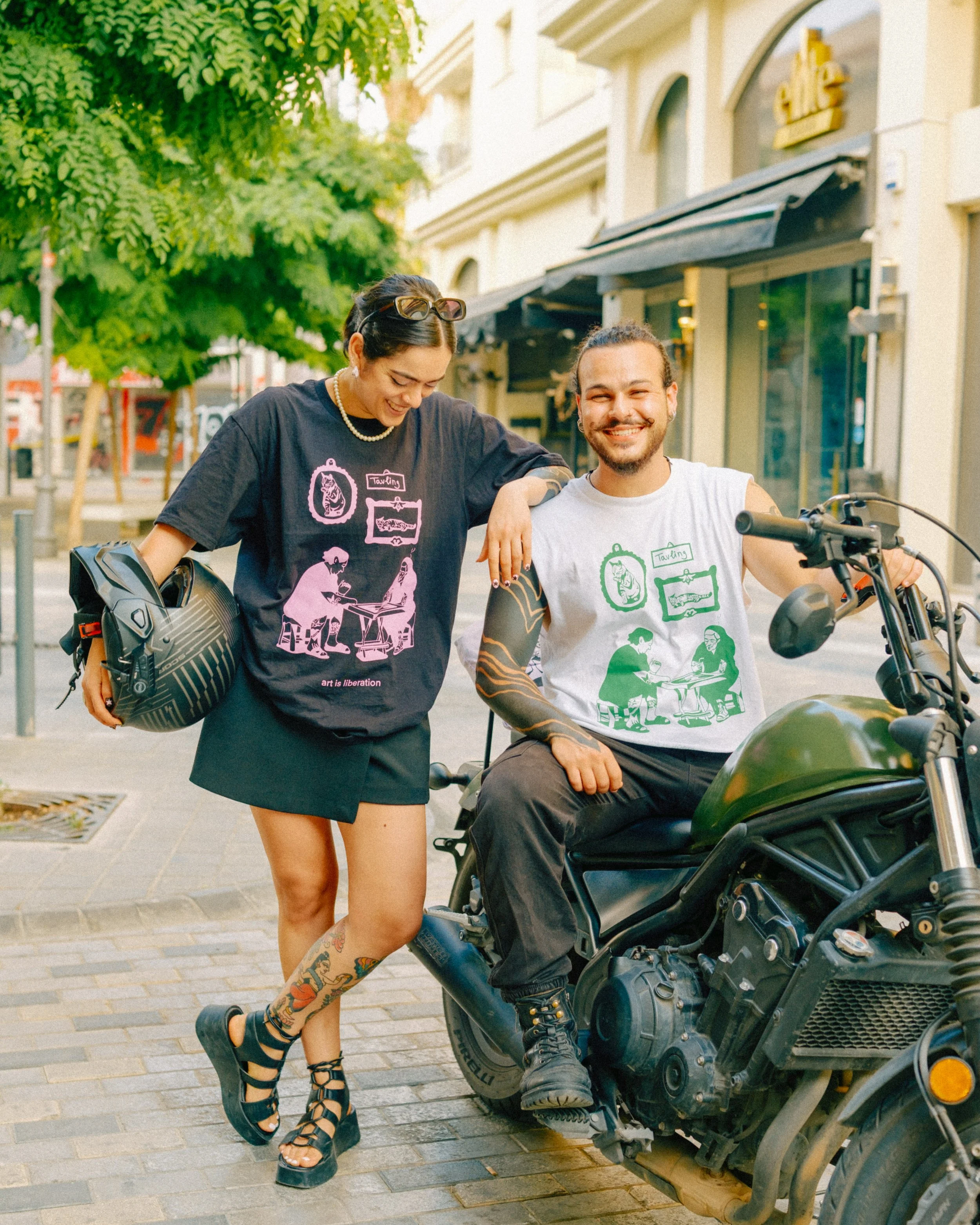 A smiling man sitting on a motorcycle and a woman standing next to him with her arm on his shoulder on a city street, both wearing matching graphic t-shirts, with green and pink outline art.