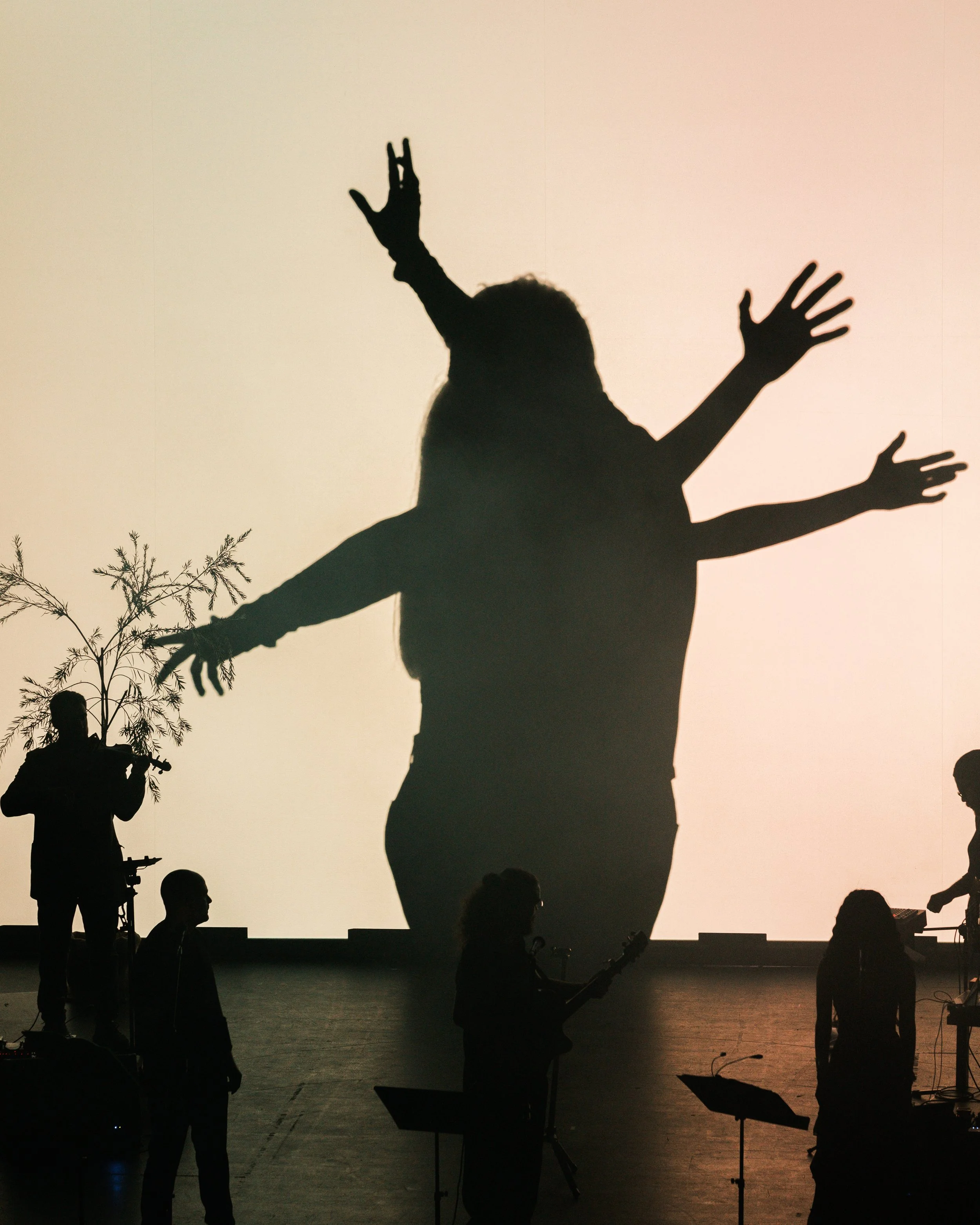 Silhouettes of a person with multiple arms in front of a large screen, with smaller musicians playing instruments and a person holding a tree branch on stage.
