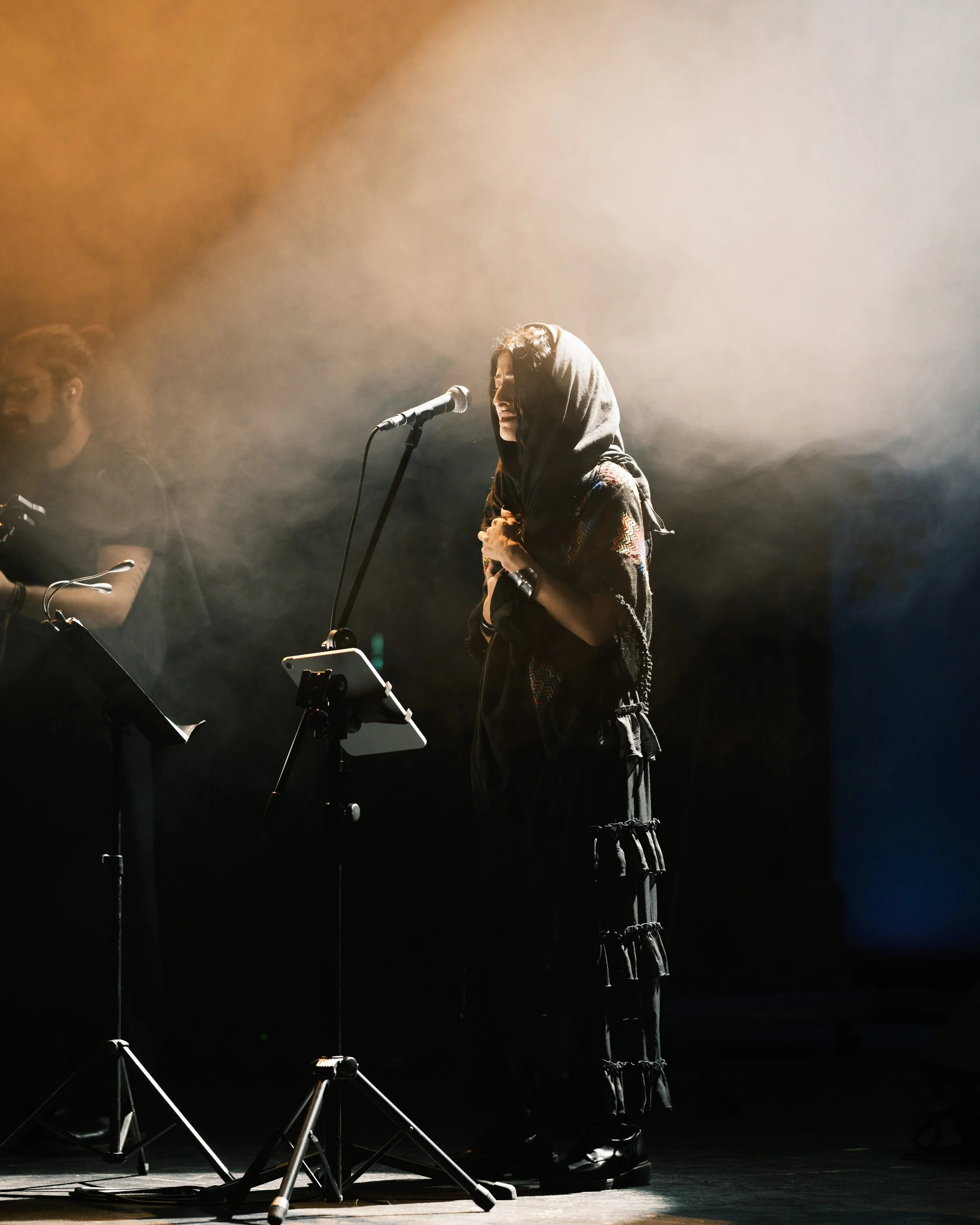 A woman singing on stage with a microphone, wearing a hooded garment and surrounded by stage lighting and fog.