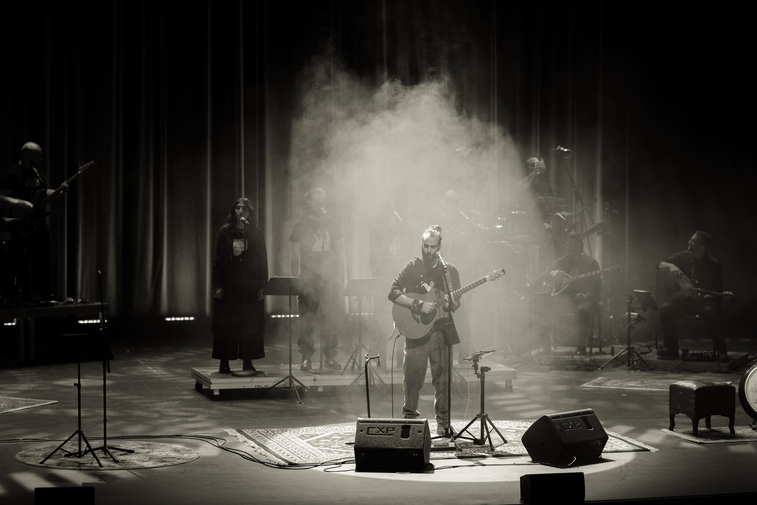 A black and white photo of a musical band performing on stage with one person in the front playing guitar and singing into a microphone, surrounded by other band members playing various instruments with stage lights and smoke effects.