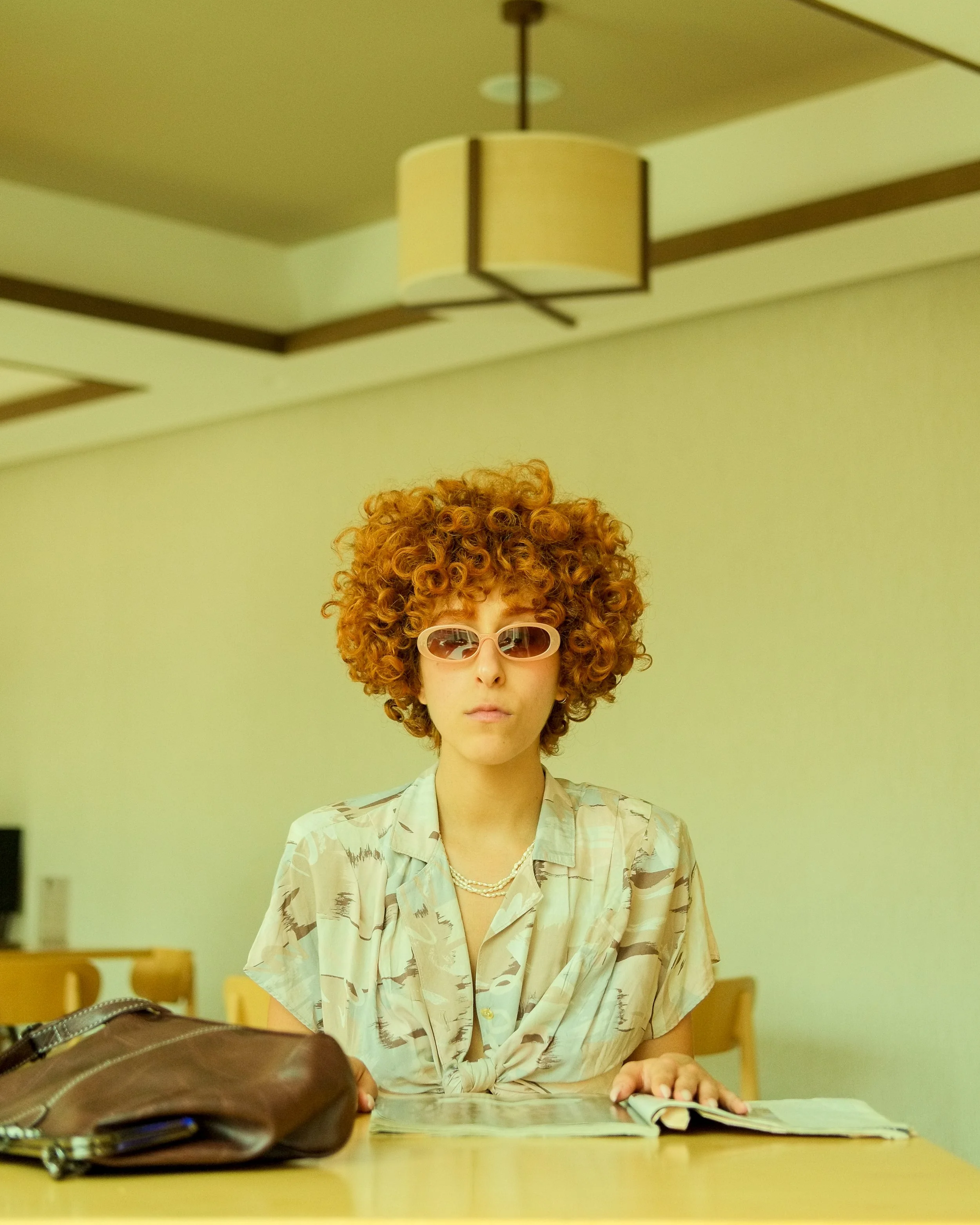 A person with curly red hair, wearing white cat-eye sunglasses, a patterned shirt, and a pearl necklace, sitting at a table with open magazines and a brown purse, in a room with yellow walls and a hanging ceiling light.