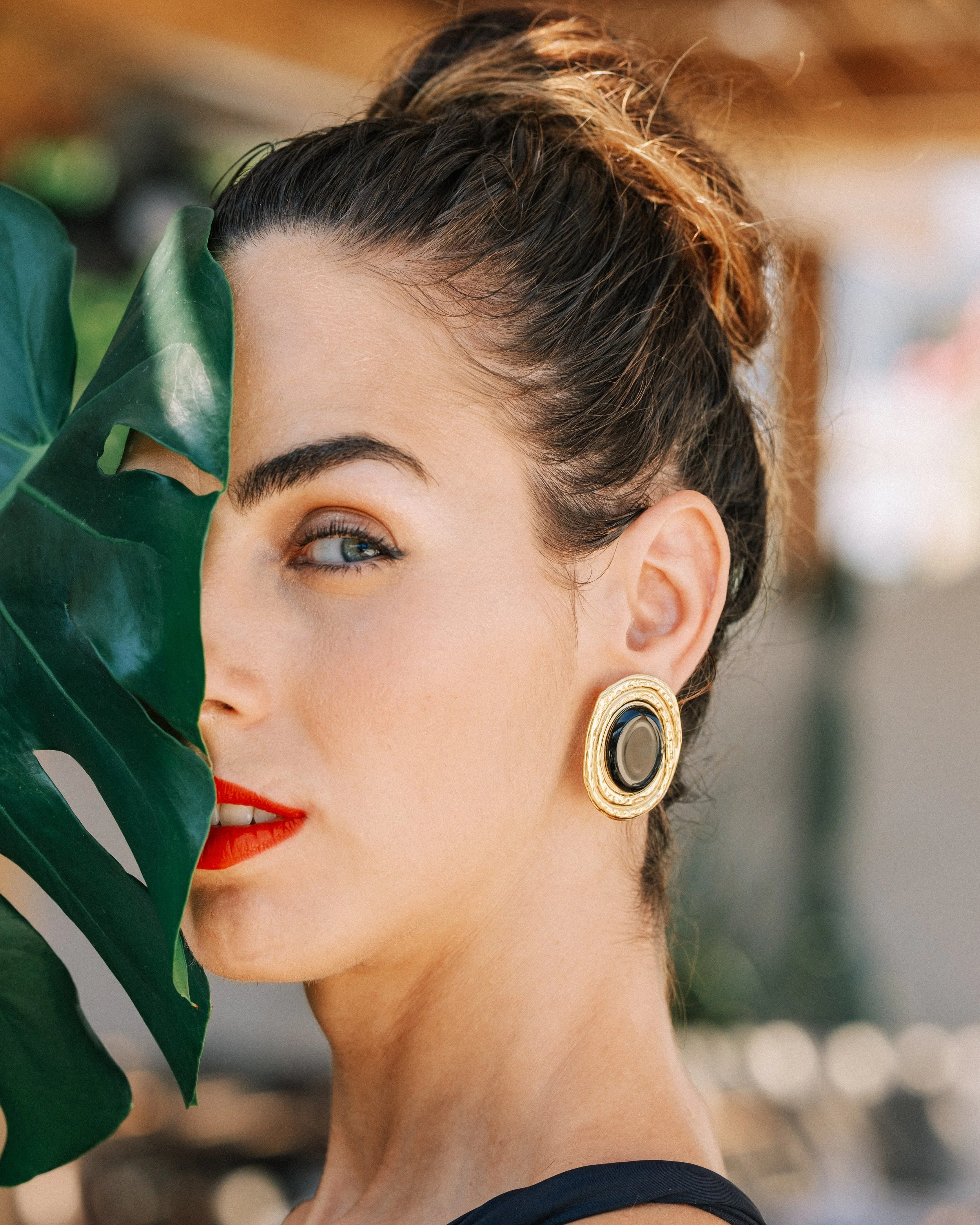 Close-up of a woman with dark hair styled in a bun, wearing earrings, with one eye visible, holding a large green tropical leaf partially covering her face, in a well-lit setting.