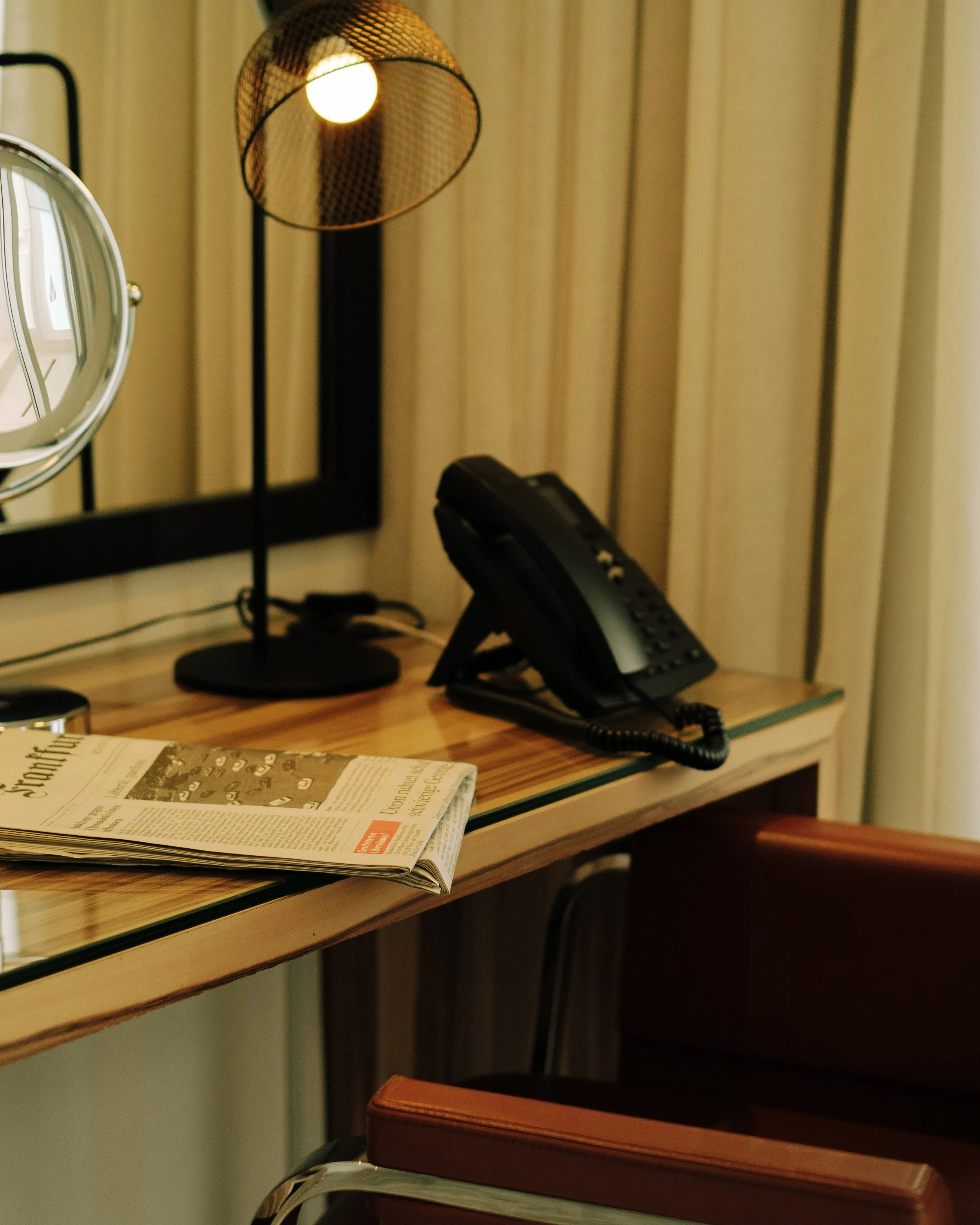 A wooden desk with a black landline phone, a standing lamp with a round shade, a mirror, a partially visible mirror, and a newspaper. There are cream-colored curtains in the background.