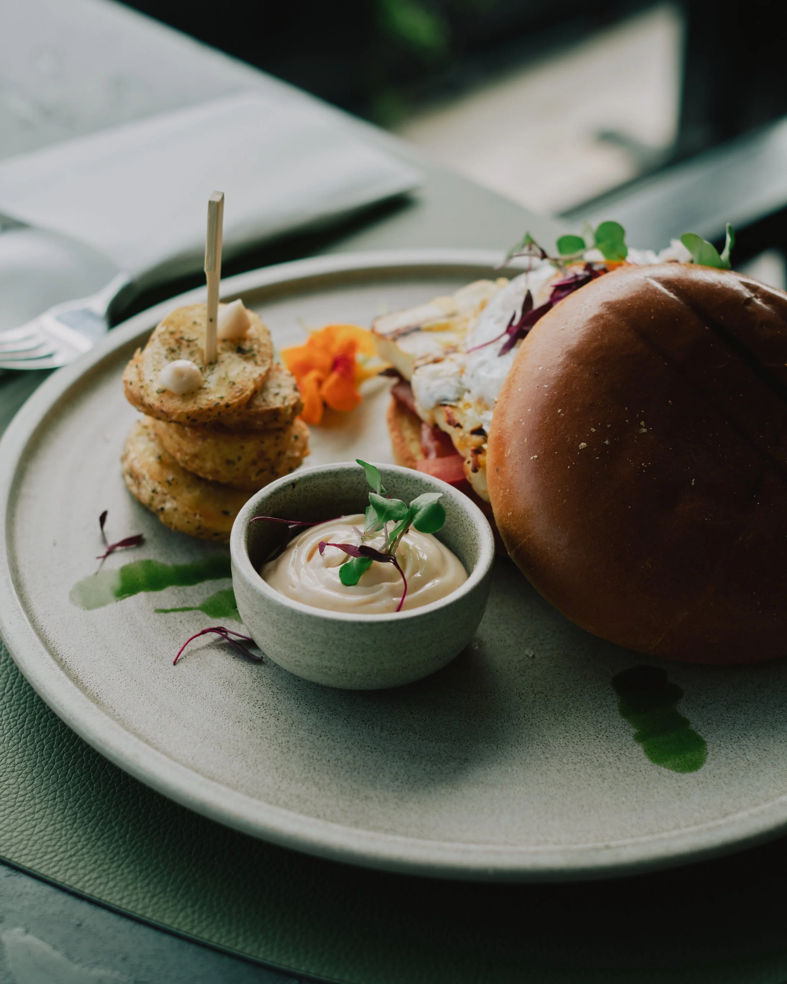 A plate with a sandwich, a small bowl of mayonnaise garnished with microgreens, and a side of potato bites, served on a ceramic plate with green sauce drizzled around.