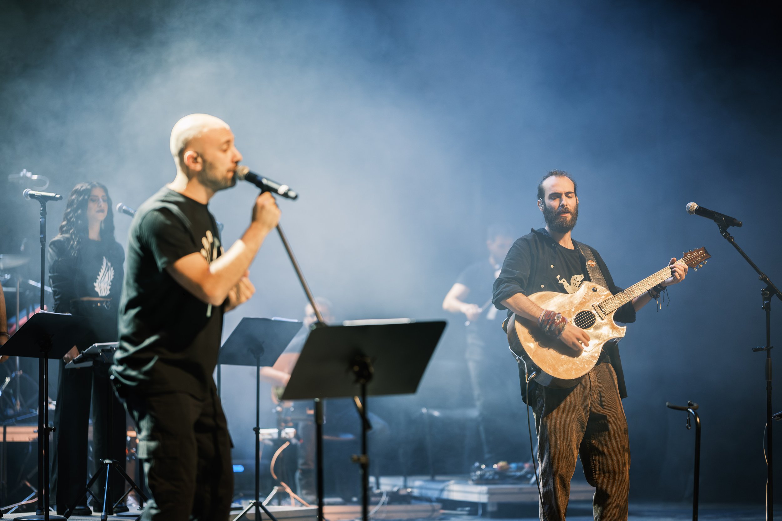 Two male musicians performing on stage with microphones and an acoustic guitar, accompanied by female musicians in the background, during a concert with concert lighting and fog effects.