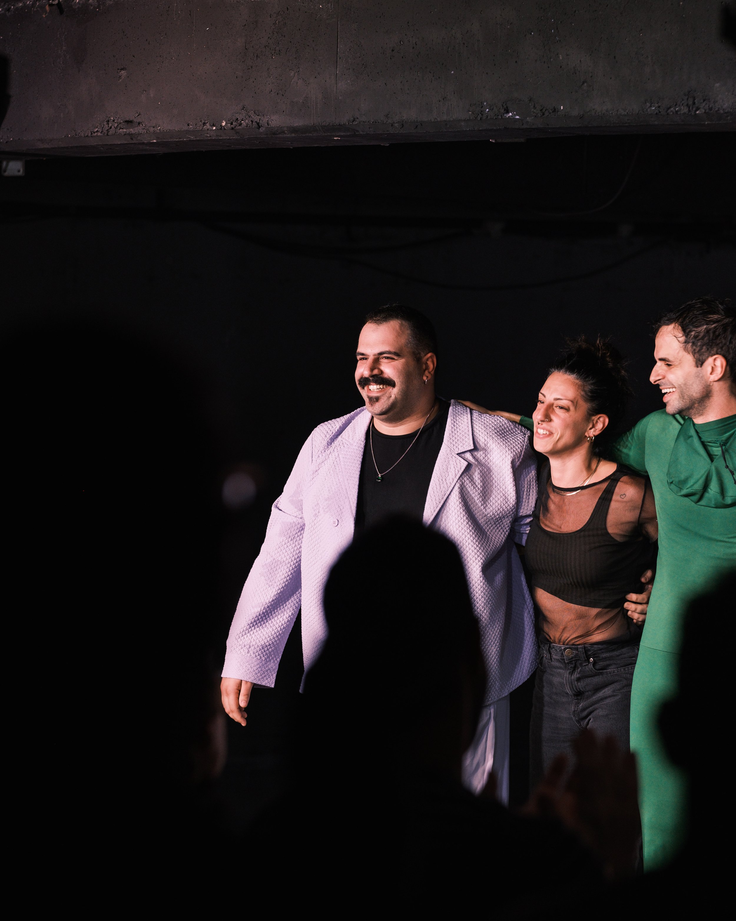 Three people standing arm-in-arm on stage, smiling, with a dark background and audience in the foreground.