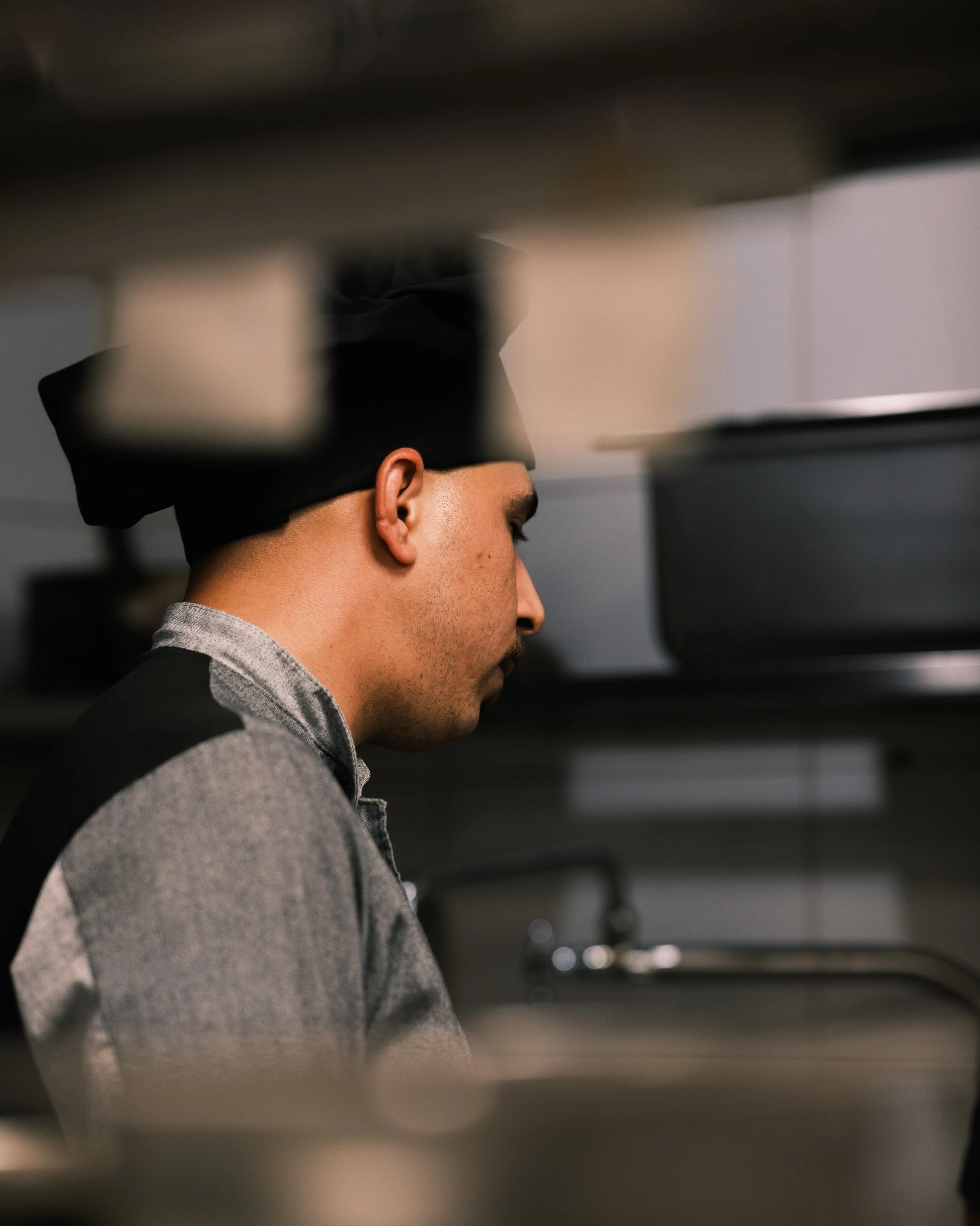 A chef in a black hat and gray uniform, seen in profile, working in a commercial kitchen.