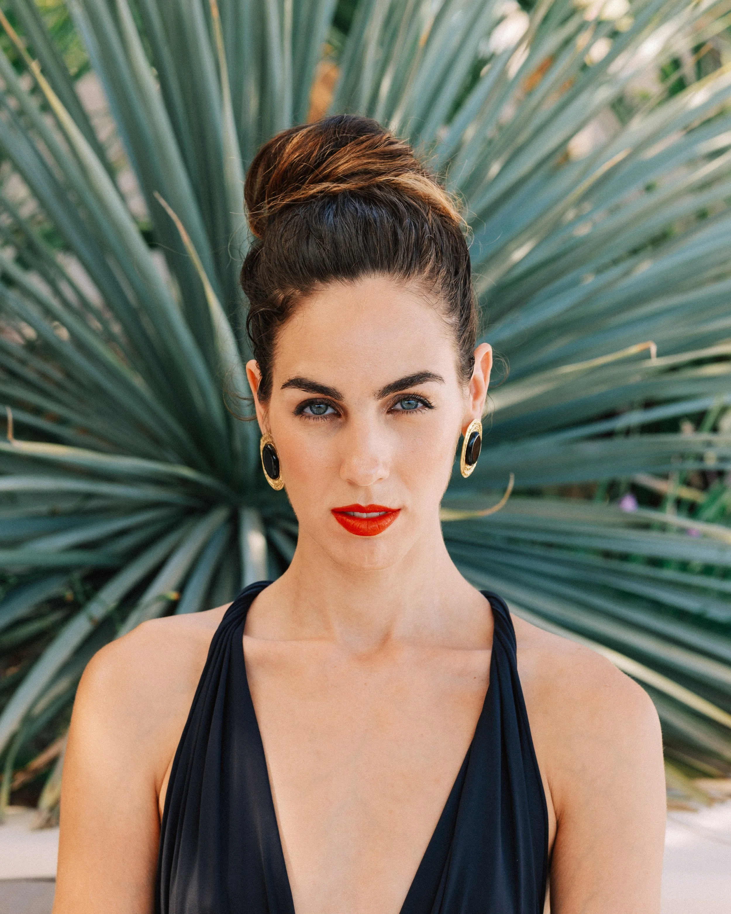 A woman with dark brown hair styled in an updo, wearing gold earrings with black stones, red lipstick, and a black sleeveless dress stands in front of a large desert plant with spiky green leaves.