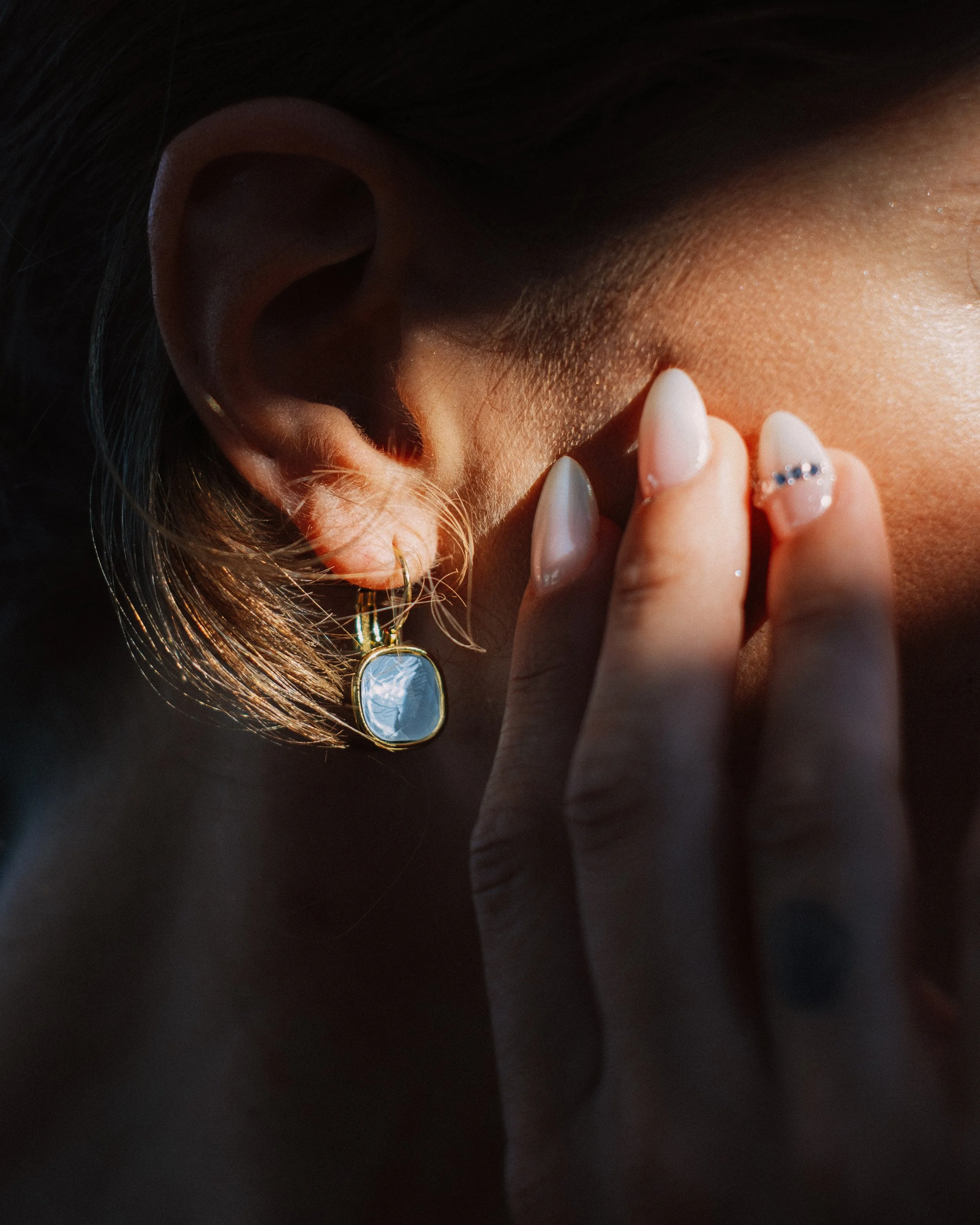 Close-up of a woman's ear with a gold earring, fingers touching her face, and her nails painted with a French manicure and rhinestone accent.