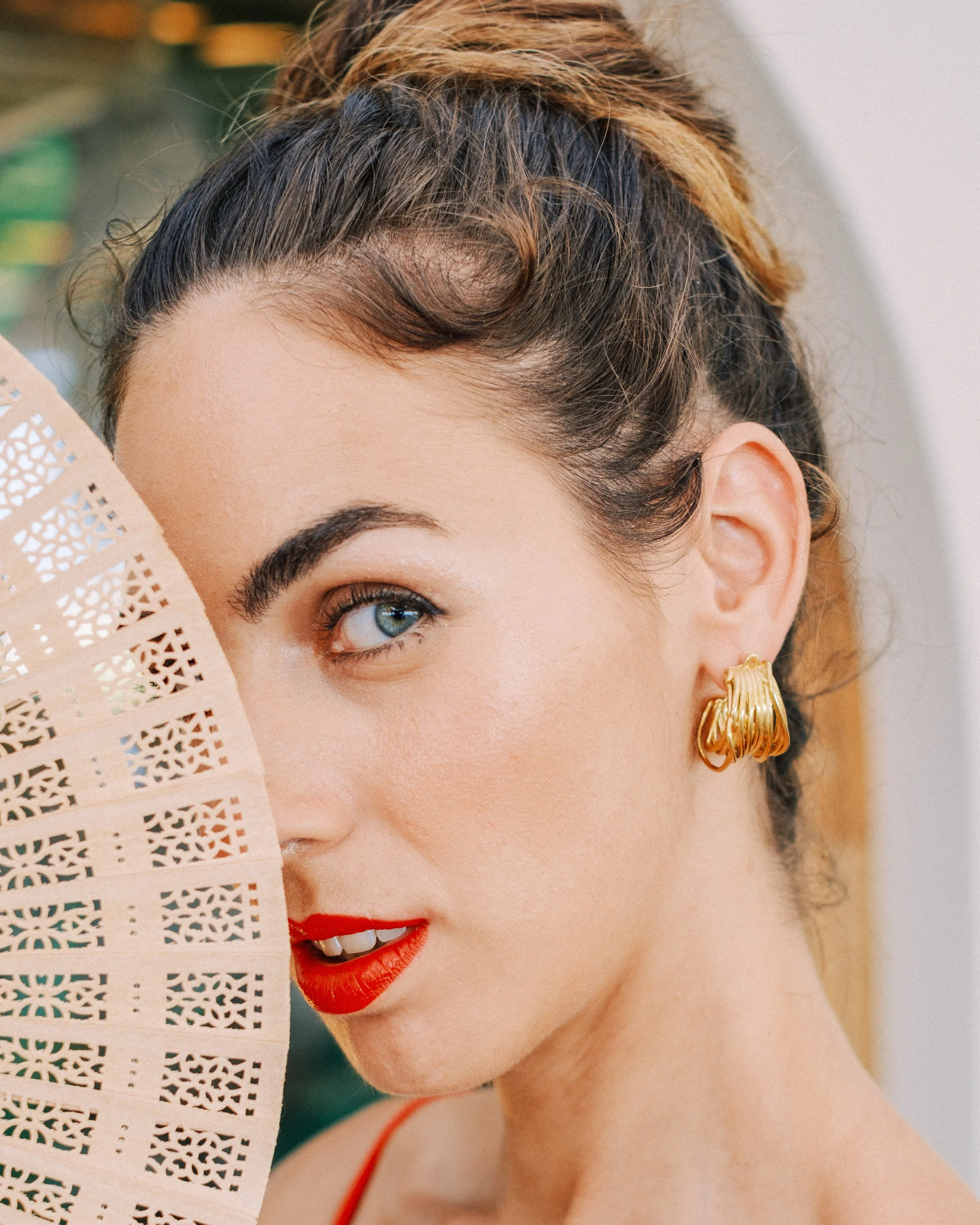 A woman with dark hair styled in an updo, wearing red lipstick and gold earrings, partially covered by a beige decorative fan.