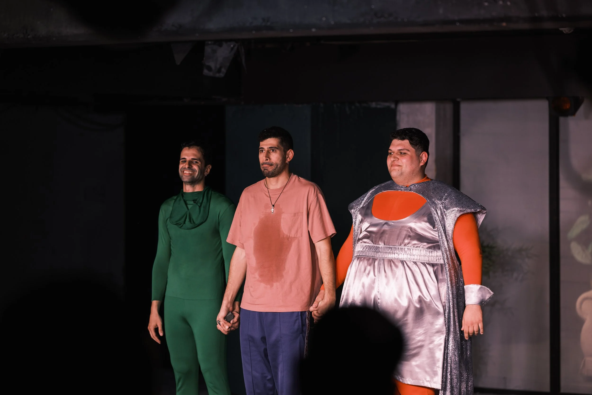 Three men standing on a stage, holding hands, with one wearing a costume with metallic silver and orange, and the other two in casual clothes.