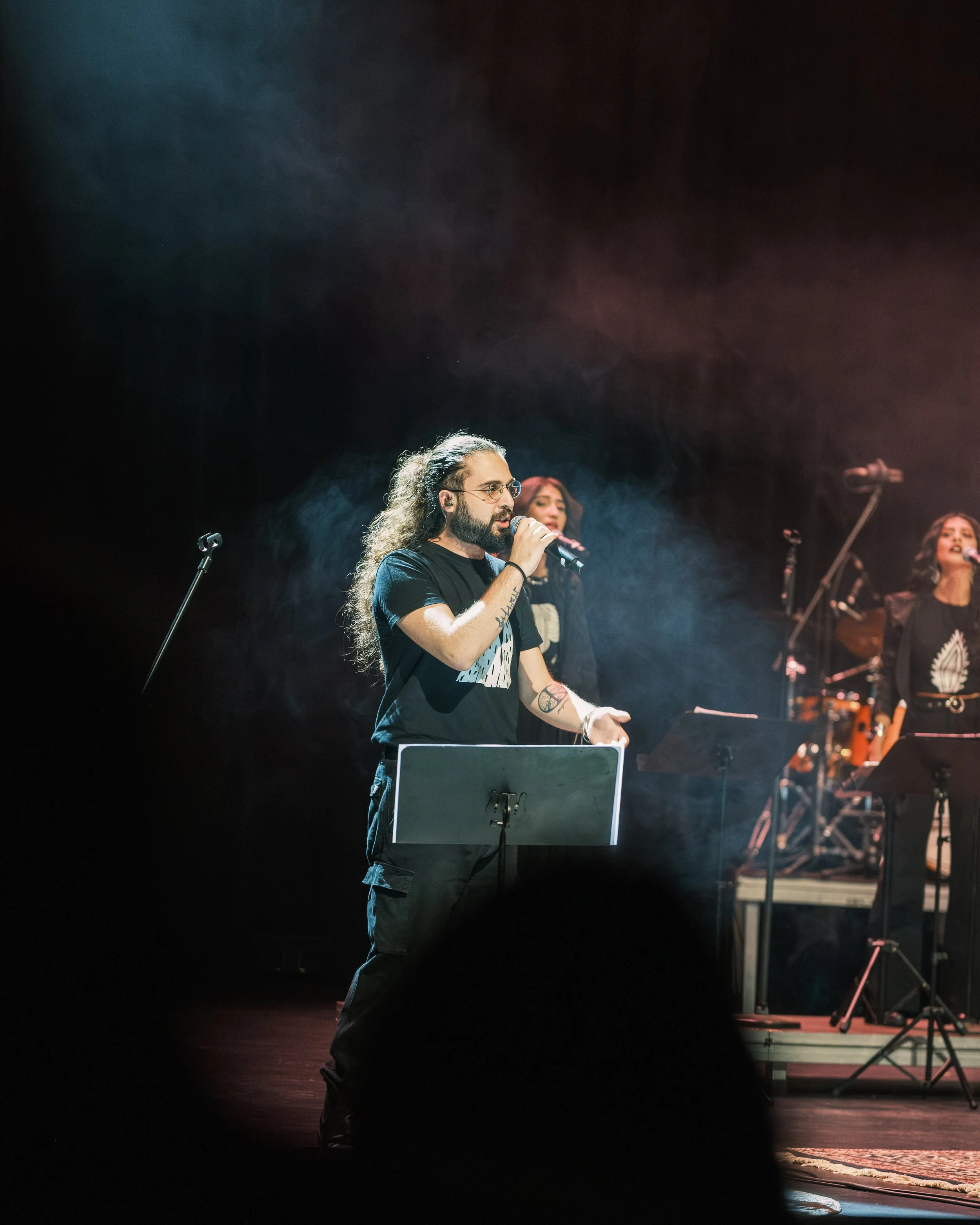 A man with long curly hair, glasses, and tattoos on his arms, holding a microphone and singing on stage with two women in the background, during a live musical performance.