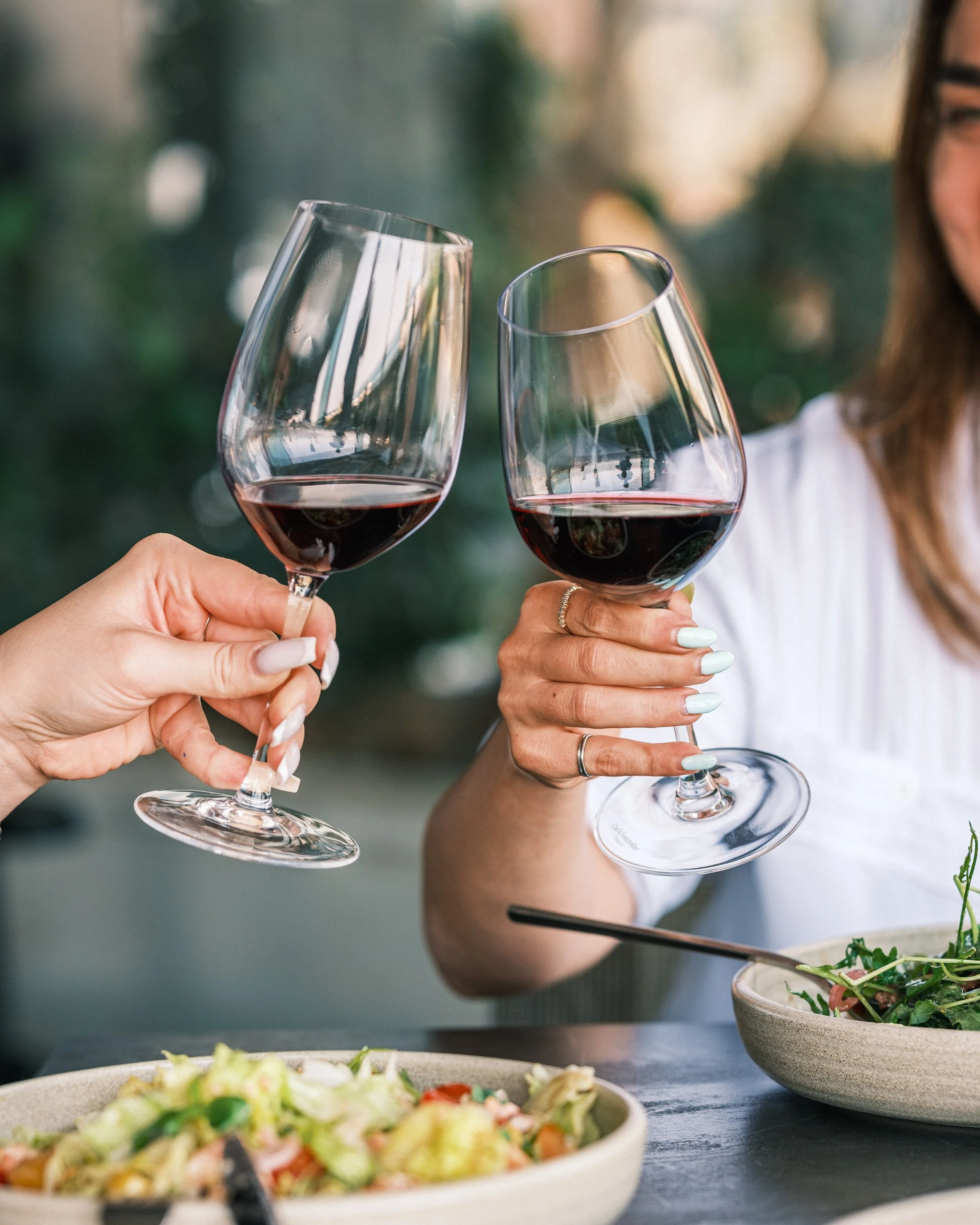 Two women toasting with glasses of red wine at a meal, with salads on the table.