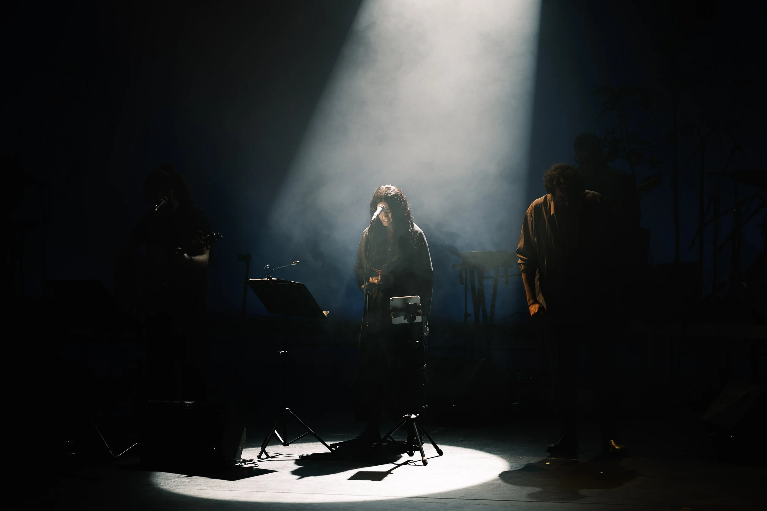 A musical performance on stage with a female singer in the center illuminated by a beam of light, flanked by band members in the dark.