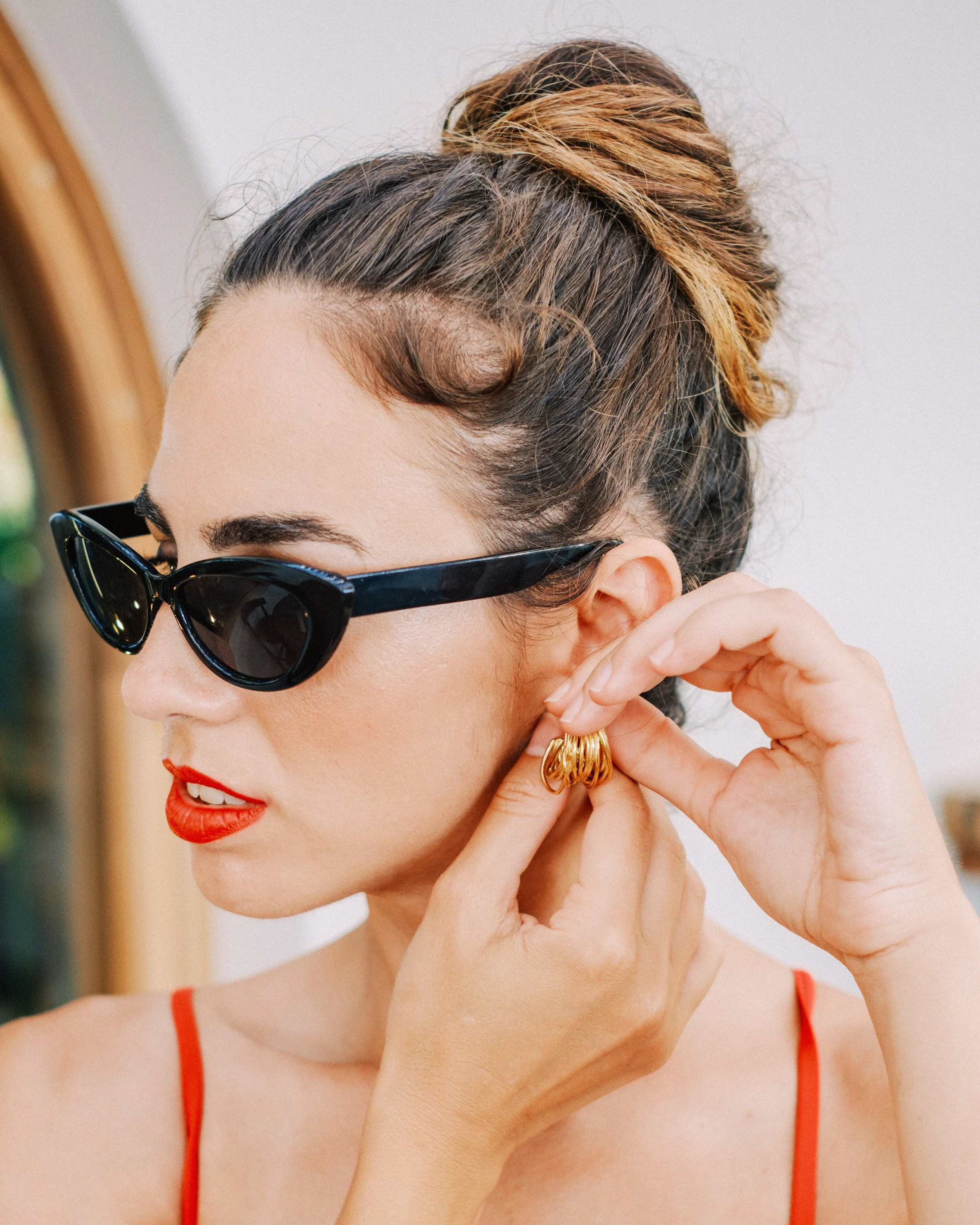 A woman with styled hair in a bun, wearing black sunglasses, red lipstick, and a red spaghetti strap top, putting on gold hoop earrings.