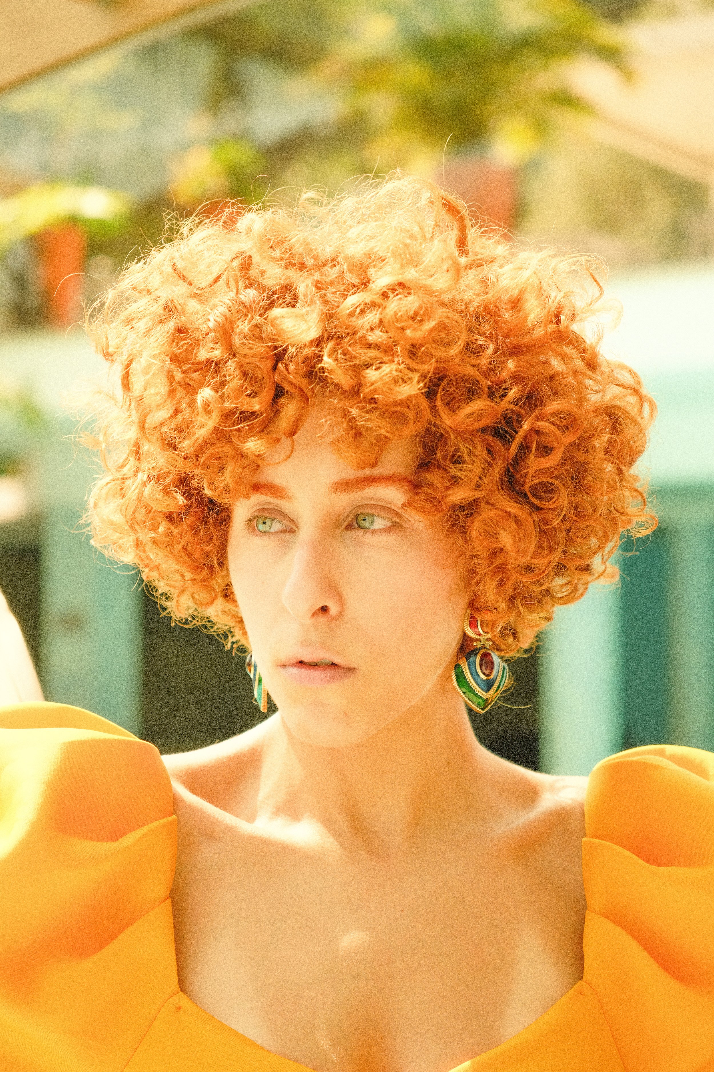 A woman with bright red, curly hair and green eyes wearing colorful earrings and an orange top with puffy sleeves.
