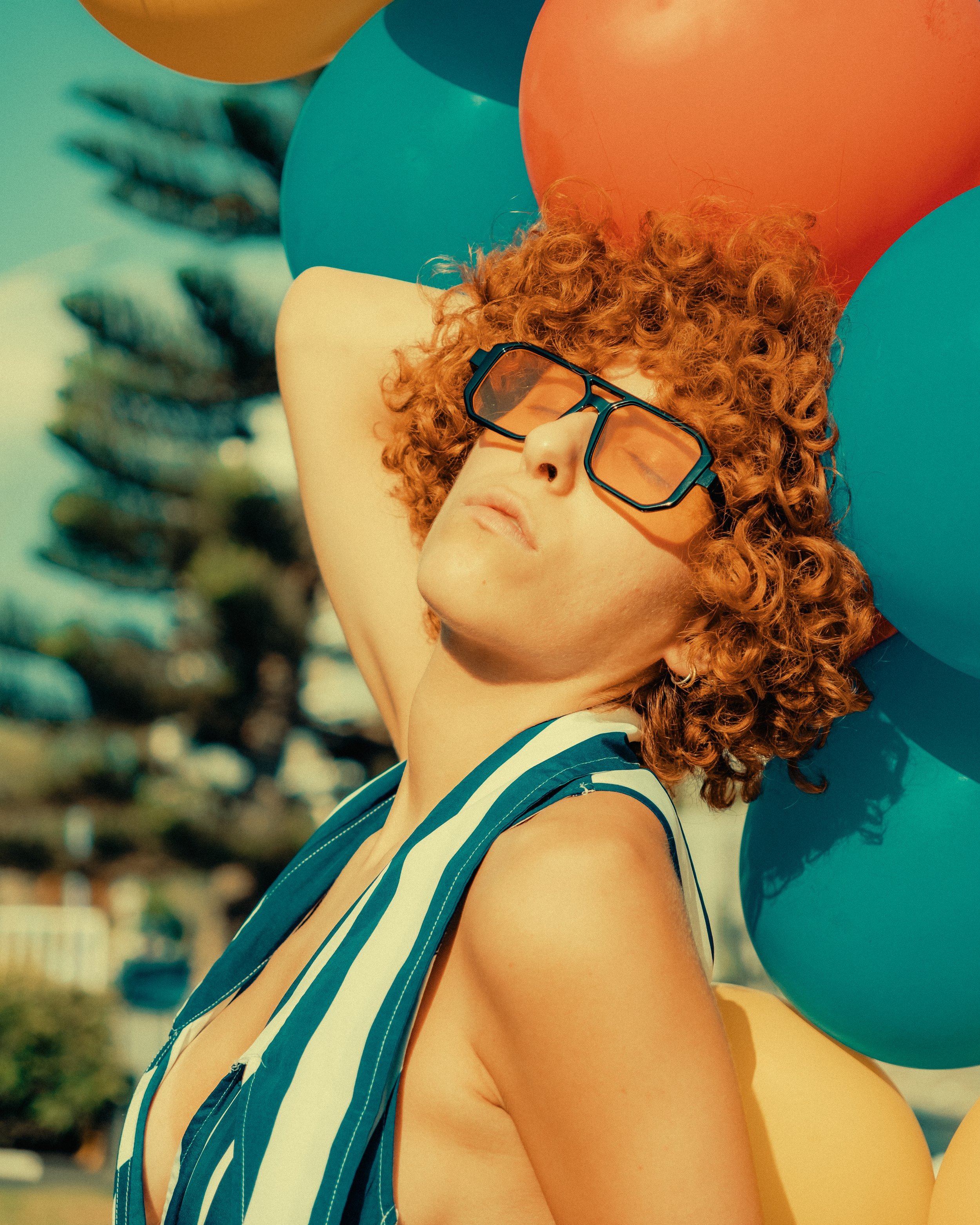 Woman with curly red hair wearing glasses and striped sleeveless top, relaxing outdoors with colorful balloons in the background.