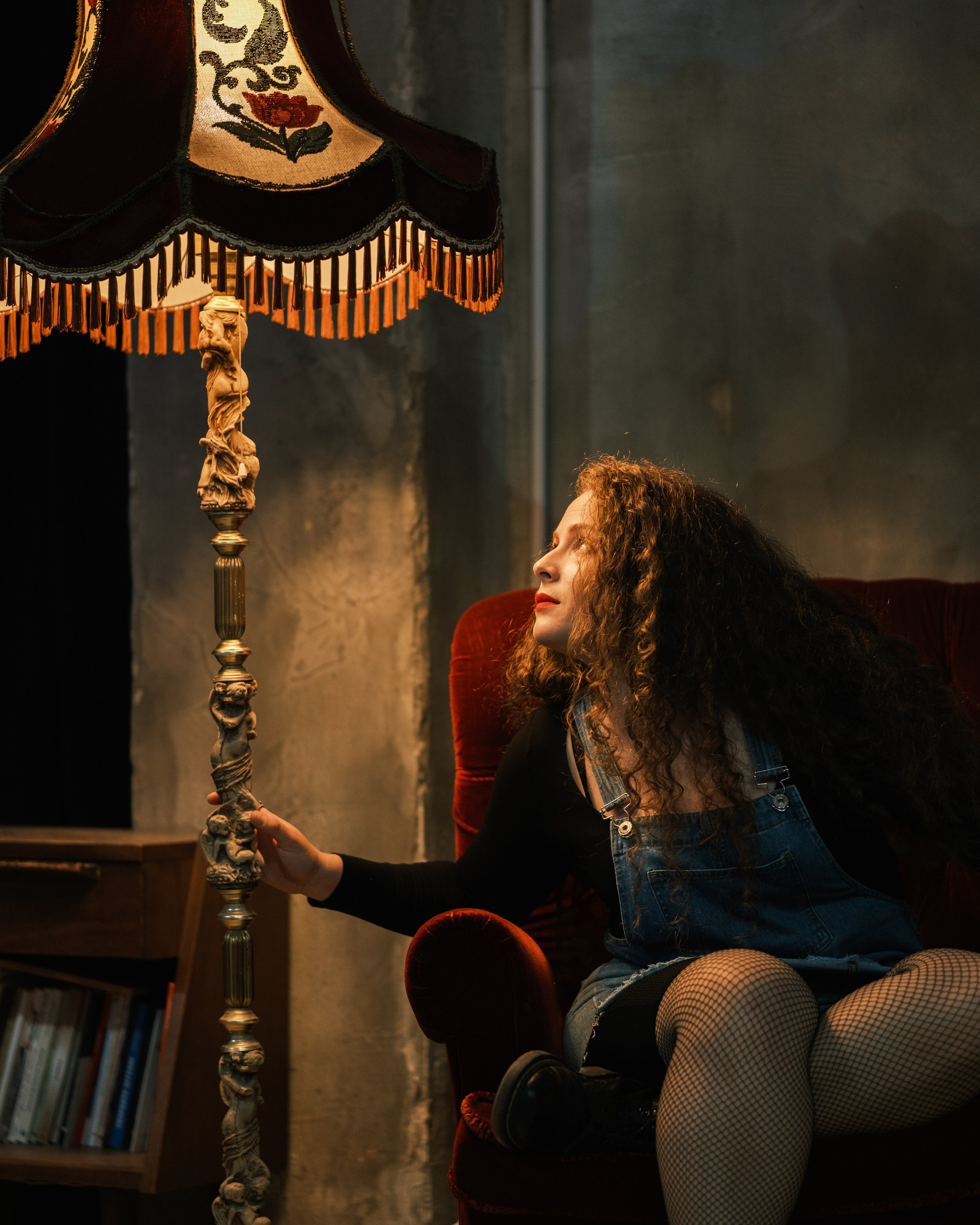 A woman with curly brown hair sitting on a red velvet armchair, looking at a vintage floor lamp with a floral lampshade, in a dimly lit room with dark walls and a bookshelf in the background.