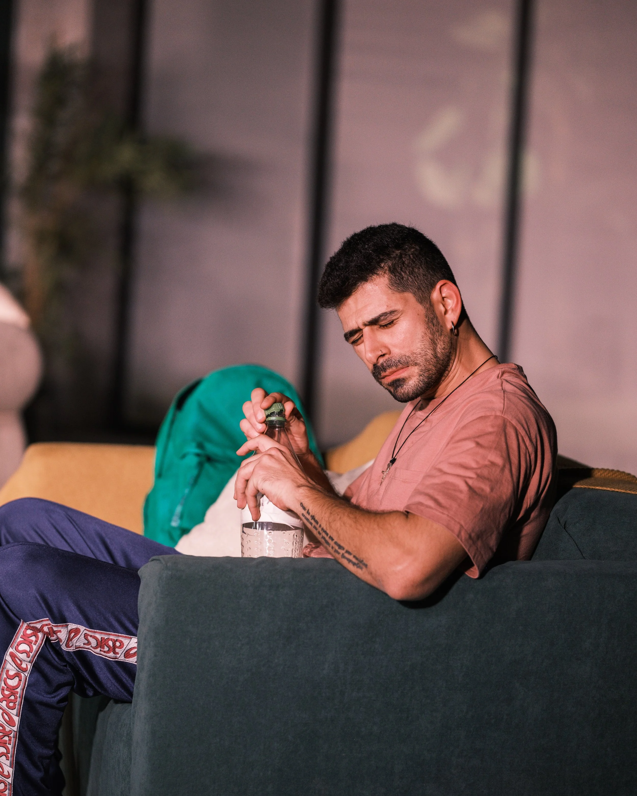 A man with a beard and tattoos, sitting on a green couch, trying to open a bottle of liquor with a concerned expression.