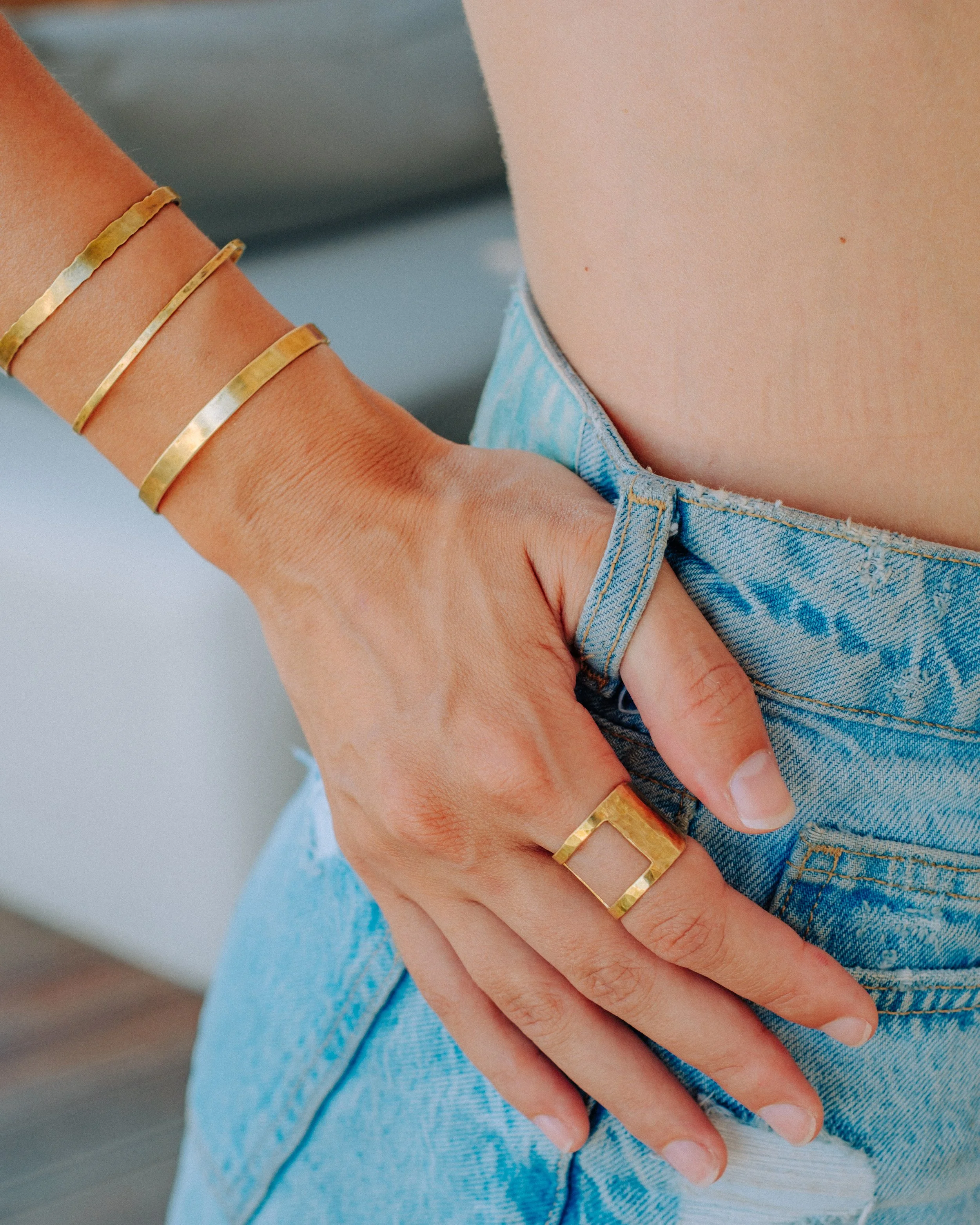 Close-up of a woman's hand resting on her hip, wearing gold jewelry including two bangles and a ring, with her thumb hooked into the belt loop of her light blue denim jeans.