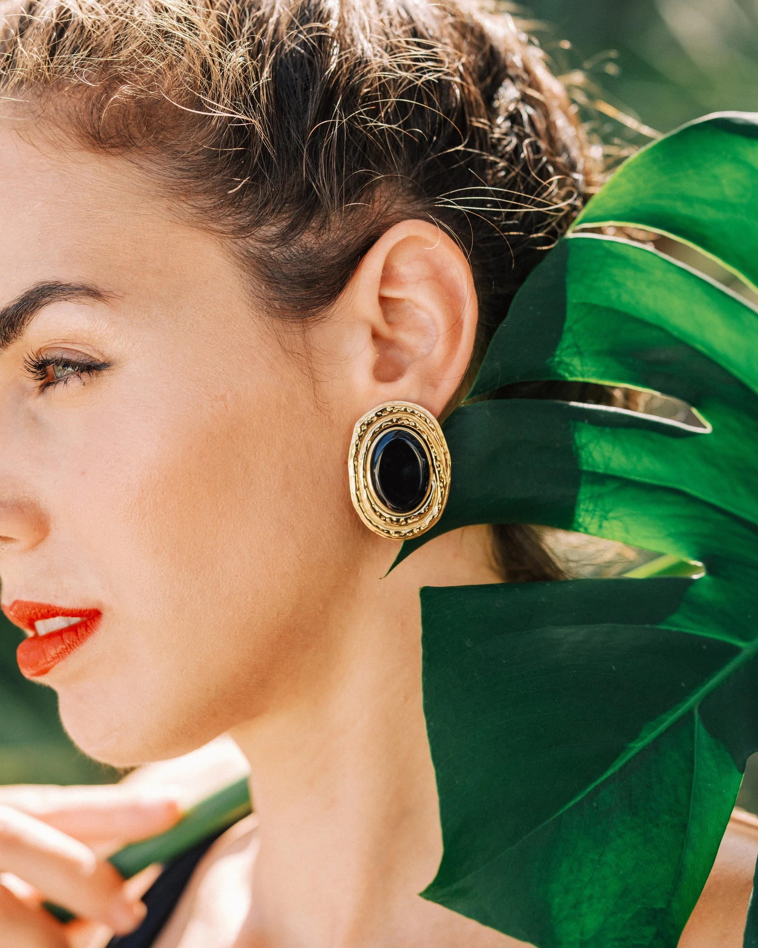 Close-up of a woman with red lipstick wearing a gold and black earring, holding a large green leaf near her face.