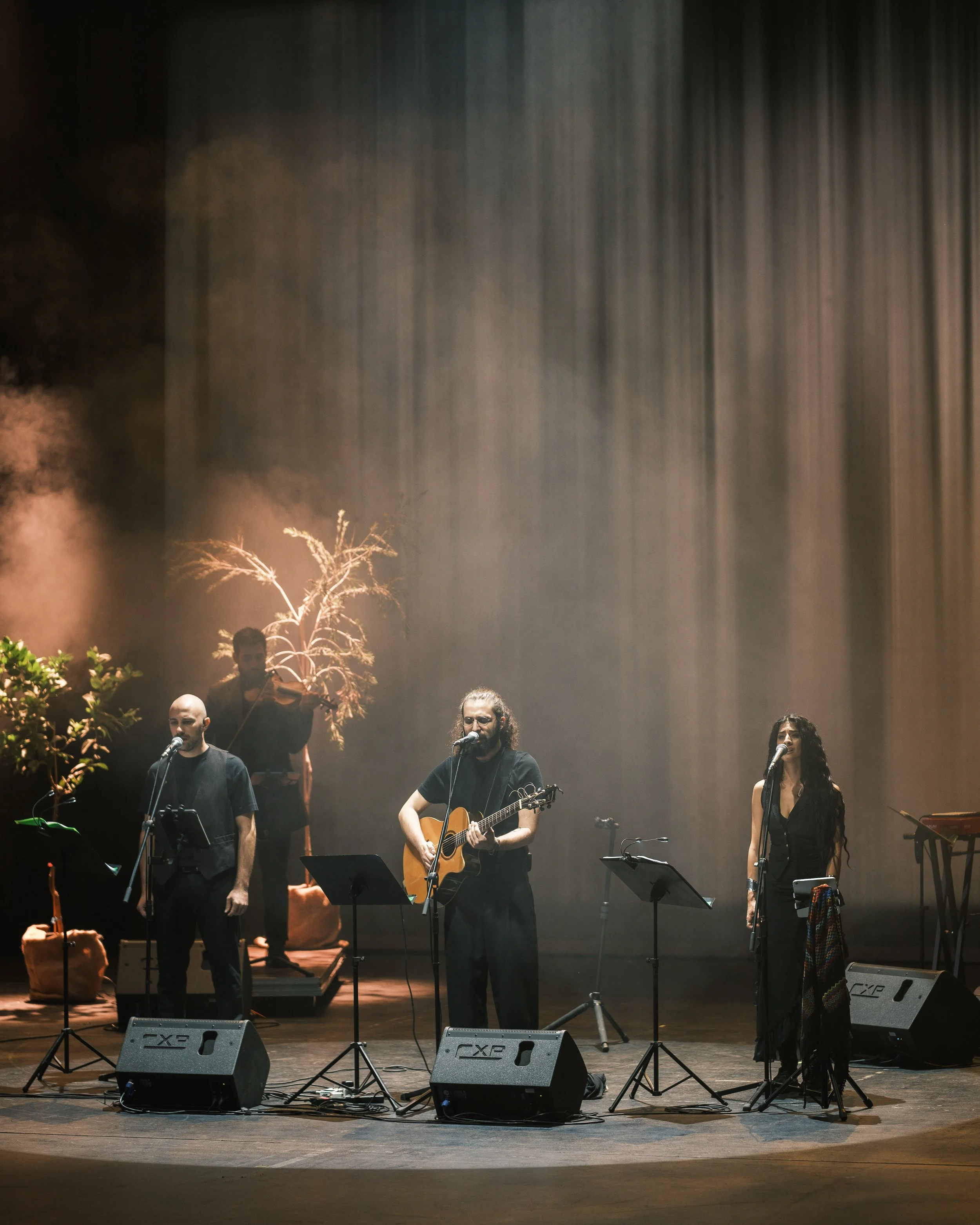 A band performing on stage with three singers at microphones and a violinist in the background, surrounded by stage props including plants and a tree.