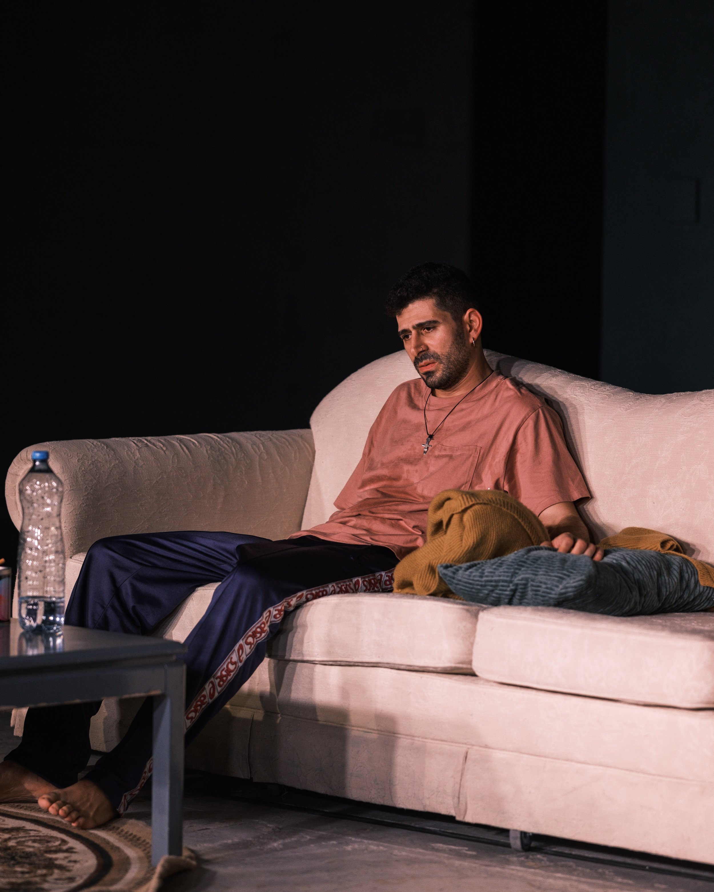 A man sitting on a beige couch with a sad or tired expression, wearing a pink shirt, navy pants, and a cross necklace, with clothes and a water bottle on the side table.