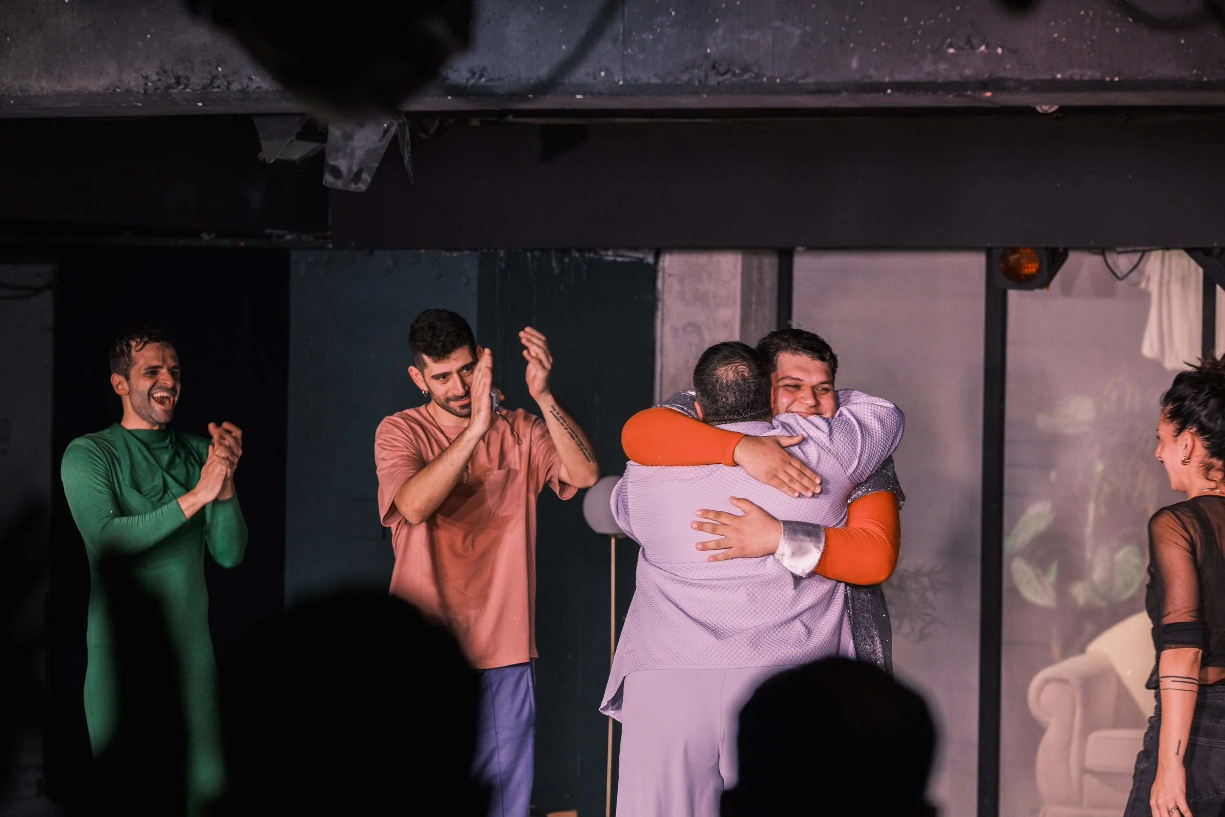 Two people hugging and smiling on stage, surrounded by three other people clapping and smiling in a theatrical or performance setting.