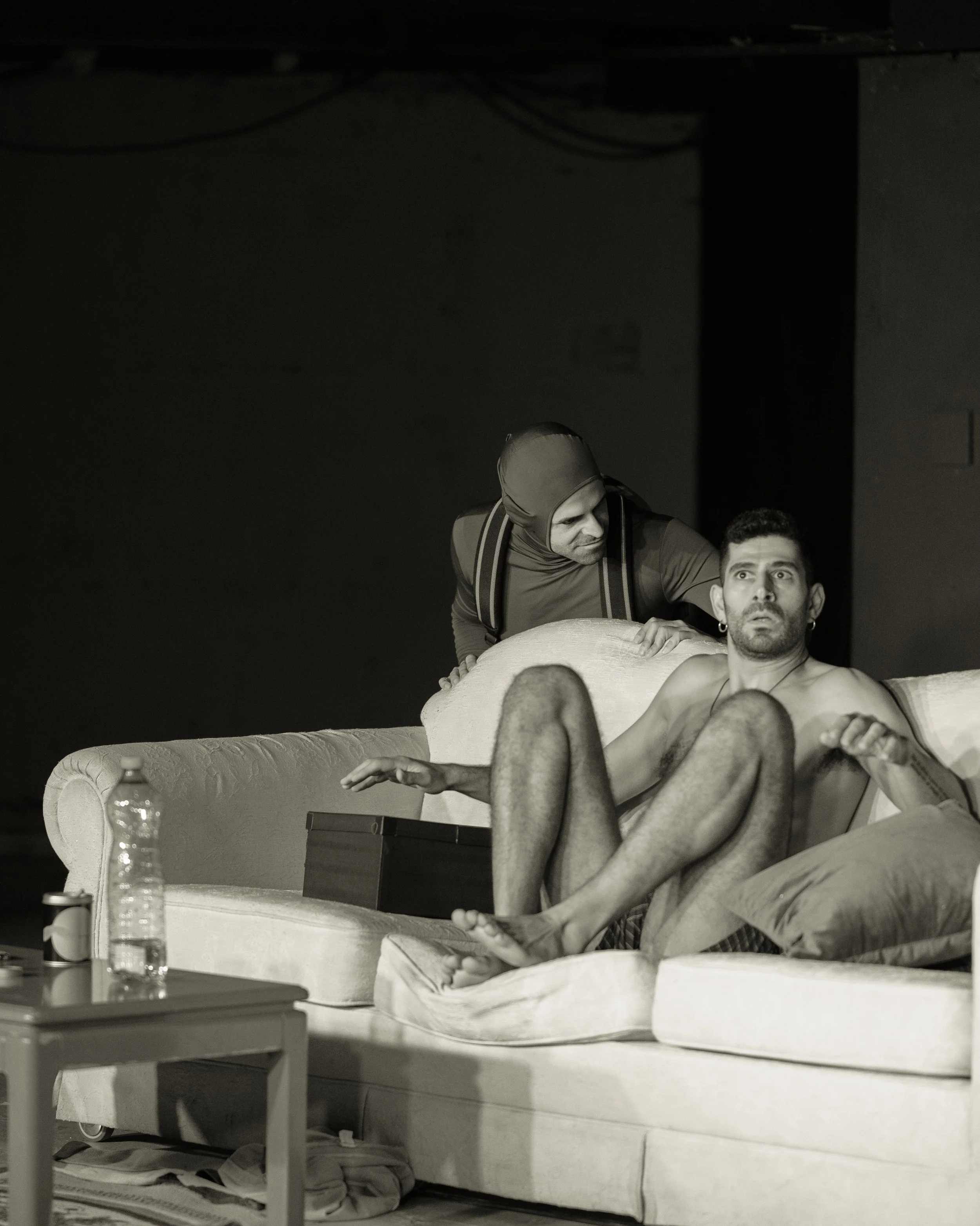 A man sitting on a sofa with a surprised expression, next to a woman, with another person standing behind, looking over him. The scene appears to be from a theatrical play or staged performance.