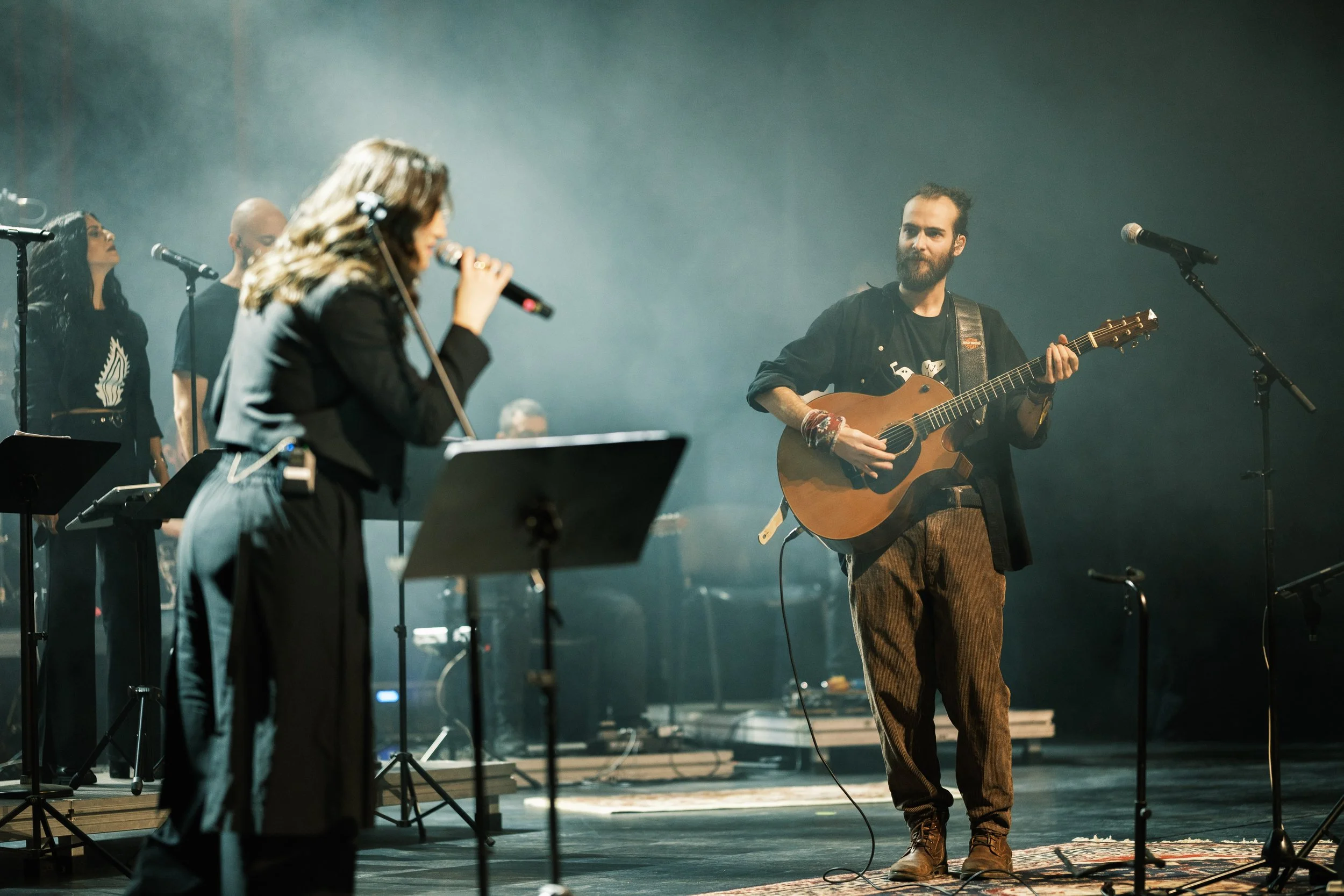 A group of musicians performing on stage, with a woman singing into a microphone and a man playing an acoustic guitar, along with other band members in the background.