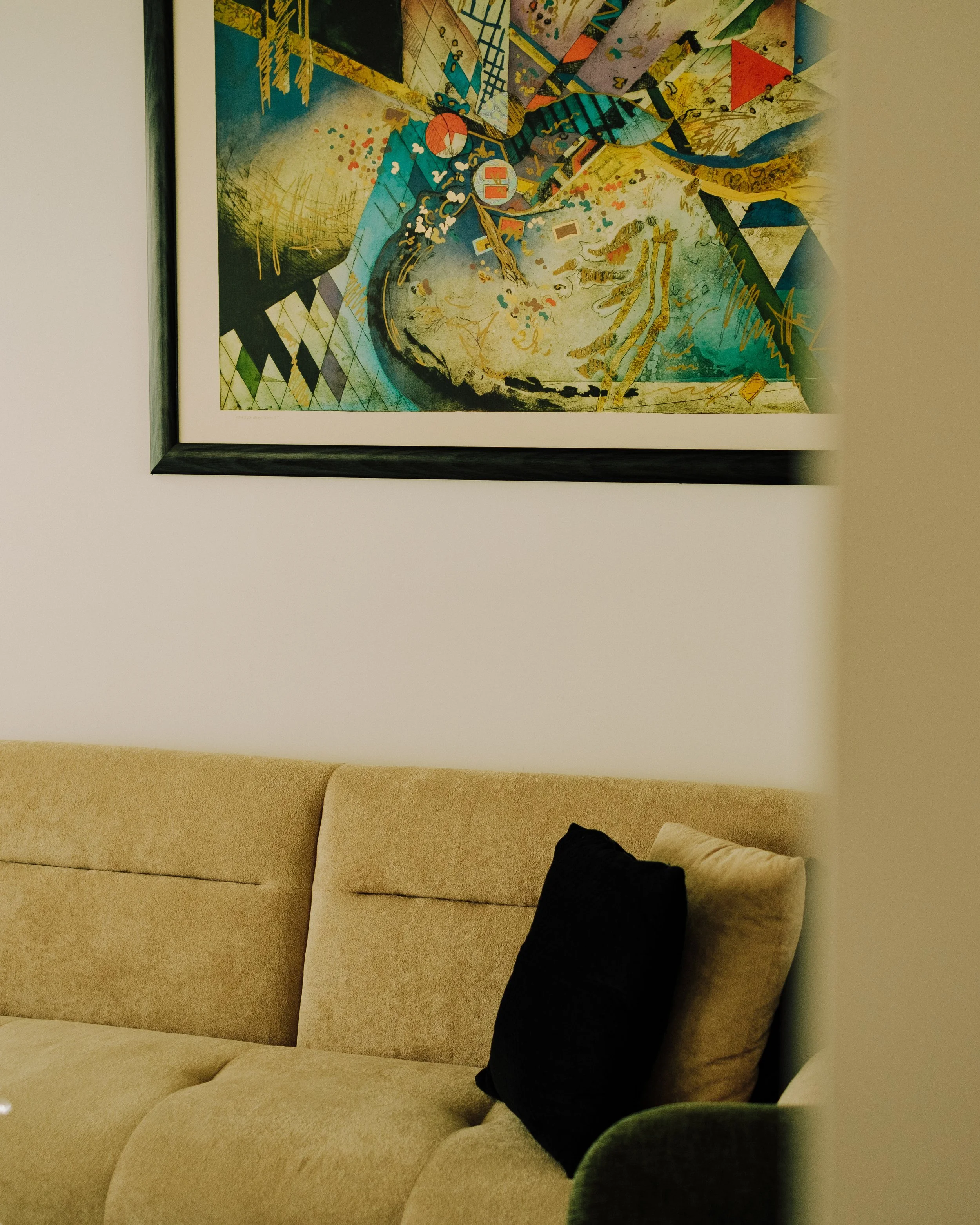 Part of a colorful abstract painting hanging on a white wall above a beige sofa with a black pillow and a beige pillow.