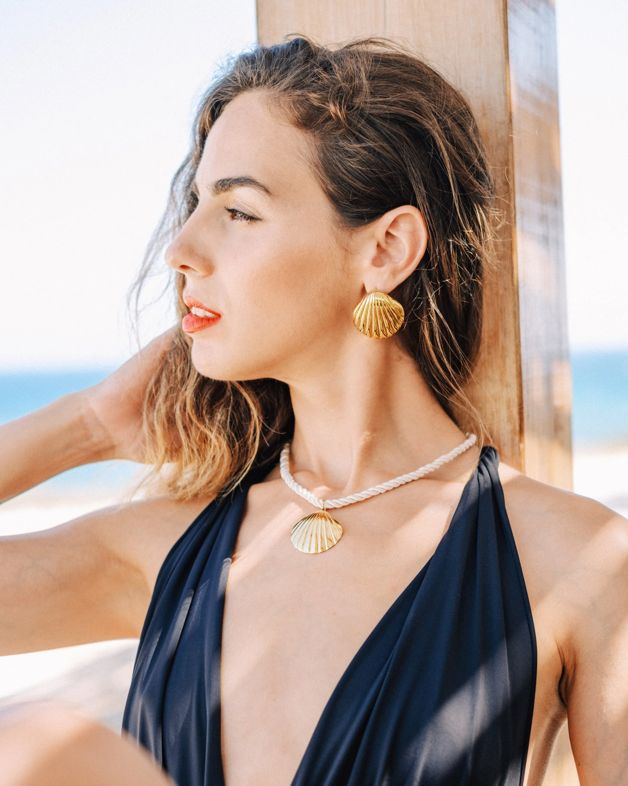 Woman with wavy brown hair wearing seashell jewelry at the beach, leaning against a wooden post