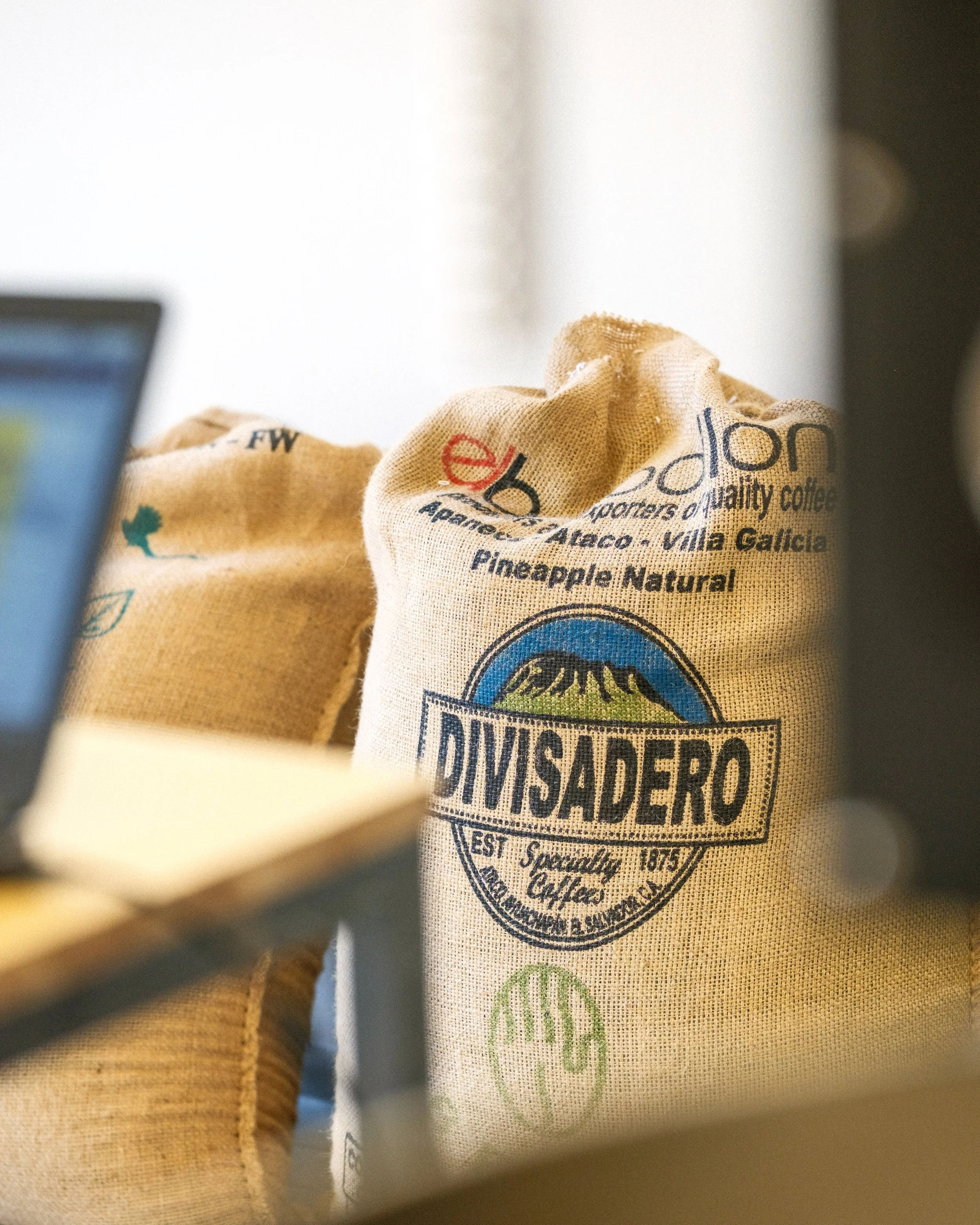 A close-up of a burlap coffee bag with 'Divisadero' logo and text, partially covering a laptop and blurred background.